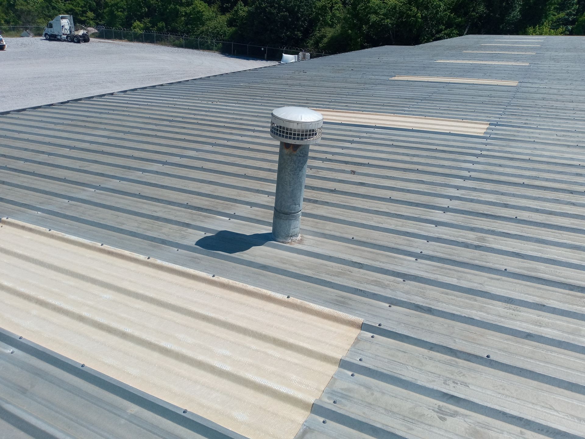 Ribbed metal roof skylights, vent pipe and light-colored repair patch under bright sunlight