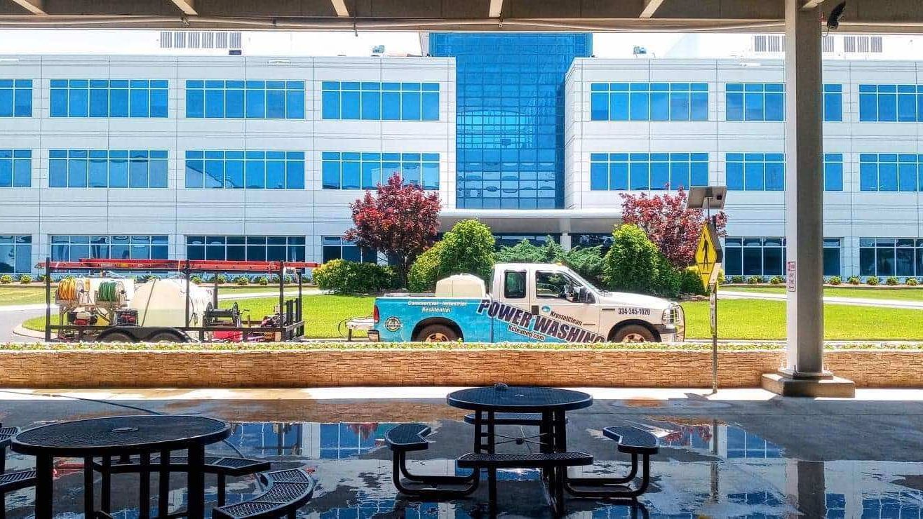 A truck is parked in front of a large building with picnic tables in front of it.
