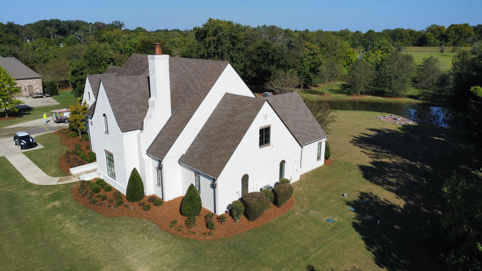Aerial view of a white, multi-gabled house with a brown roof, surrounded by green lawns and trees near a small pond.