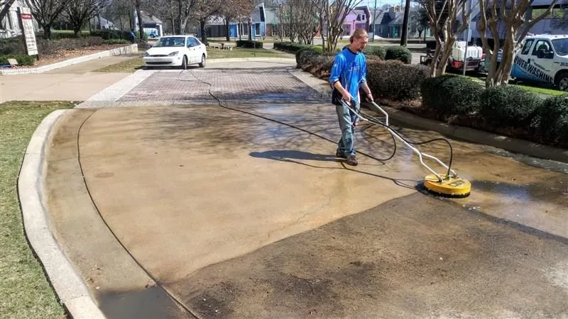 Worker using a surface cleaner to pressure wash a stained concrete driveway.