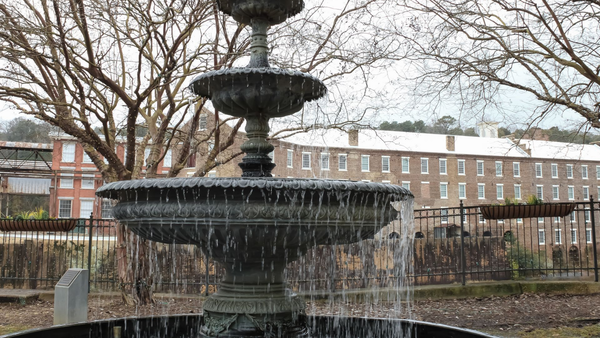 A tiered, dark metal fountain spraying water in a public park with bare trees and a brick building in the background.