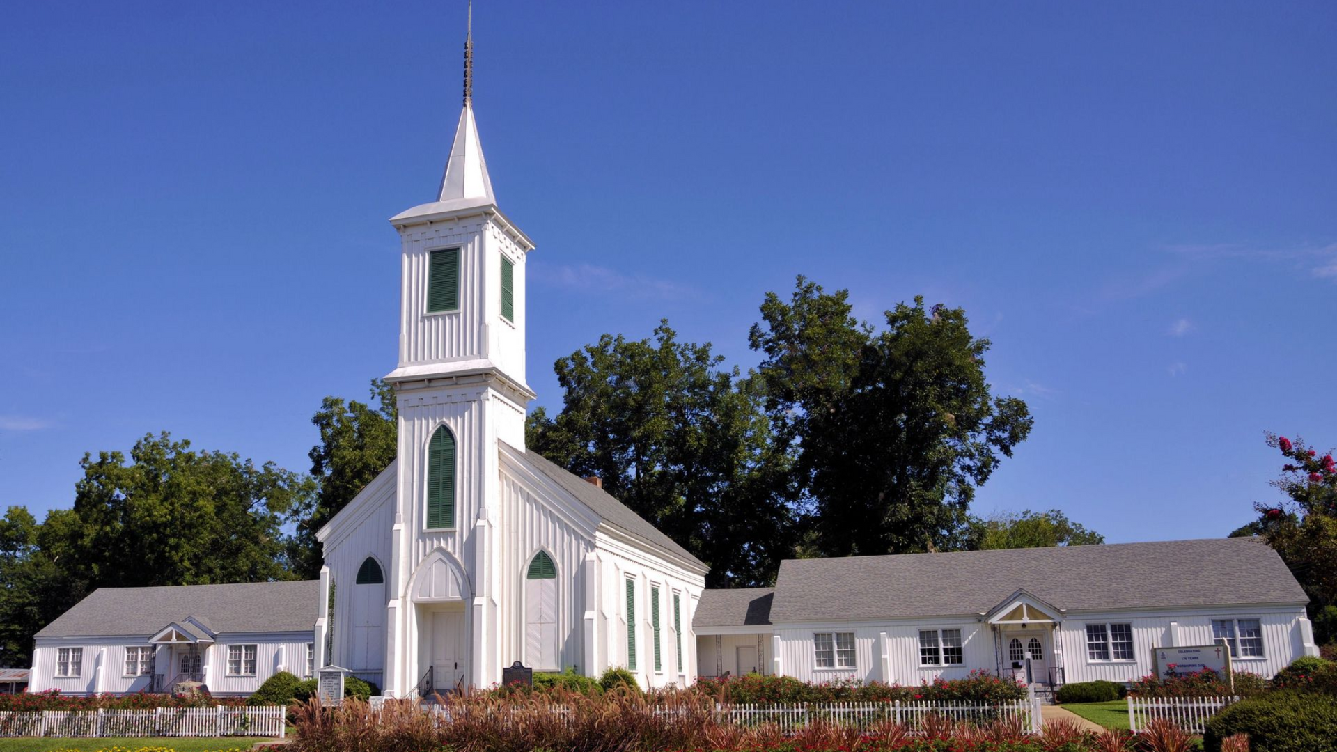 White church with steeple and green accents in Wetumpka Alabama against a blue sky, surrounded by trees and shrubs.