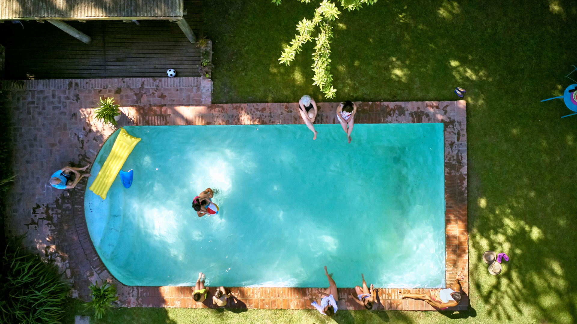 Aerial view of a backyard pool with people swimming and relaxing on the deck, surrounded by grass and trees