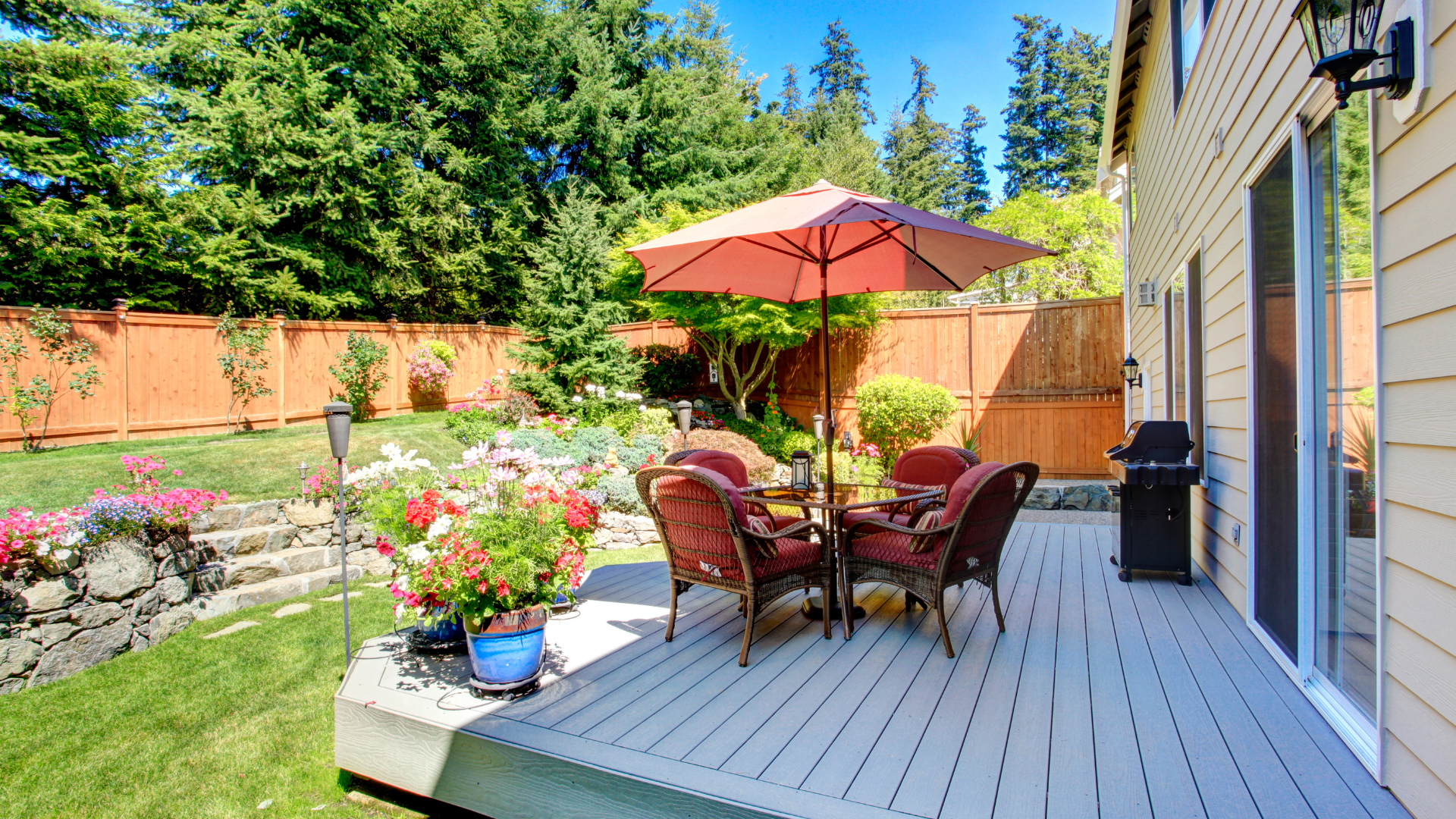 Backyard patio with wicker table set, red umbrella, potted flowers, and grill beside a house