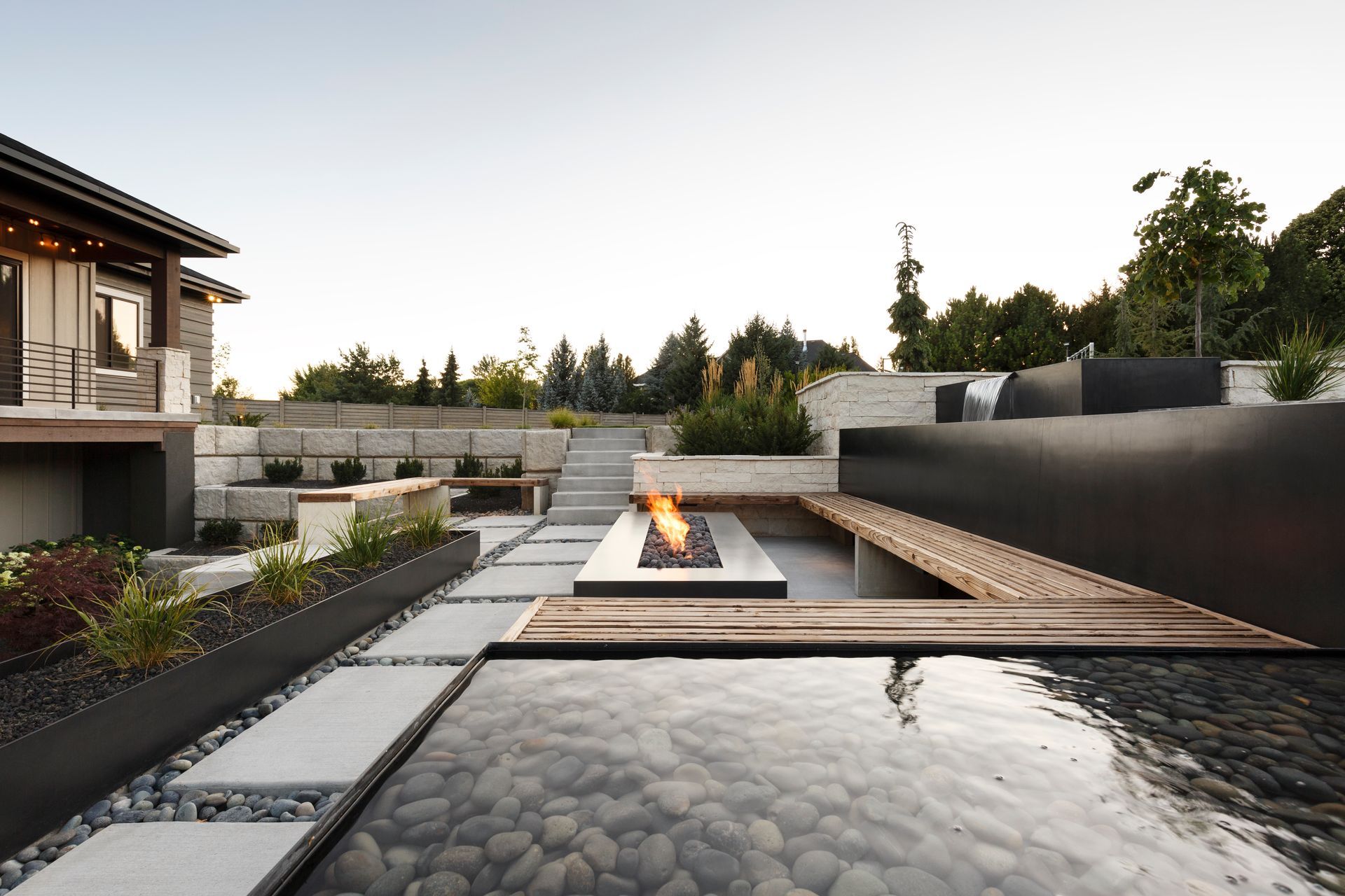 A tiered stone patio with a water feature, lounge chair, gas fire, and landscaped flower pots at dusk.