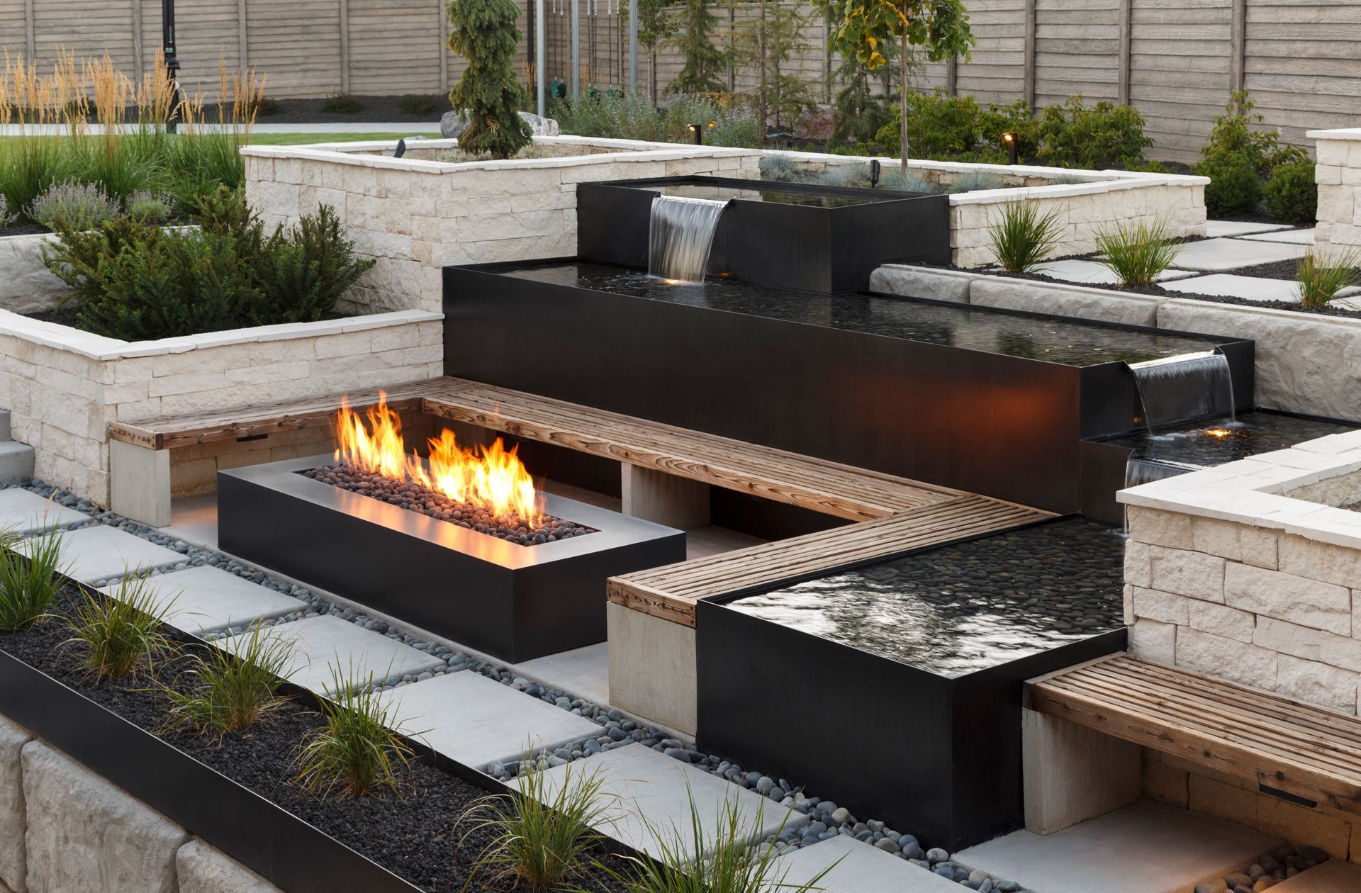A stone patio with a rectangular swimming pool, a water feature, a waterslide tunnel, and tiered landscaping.