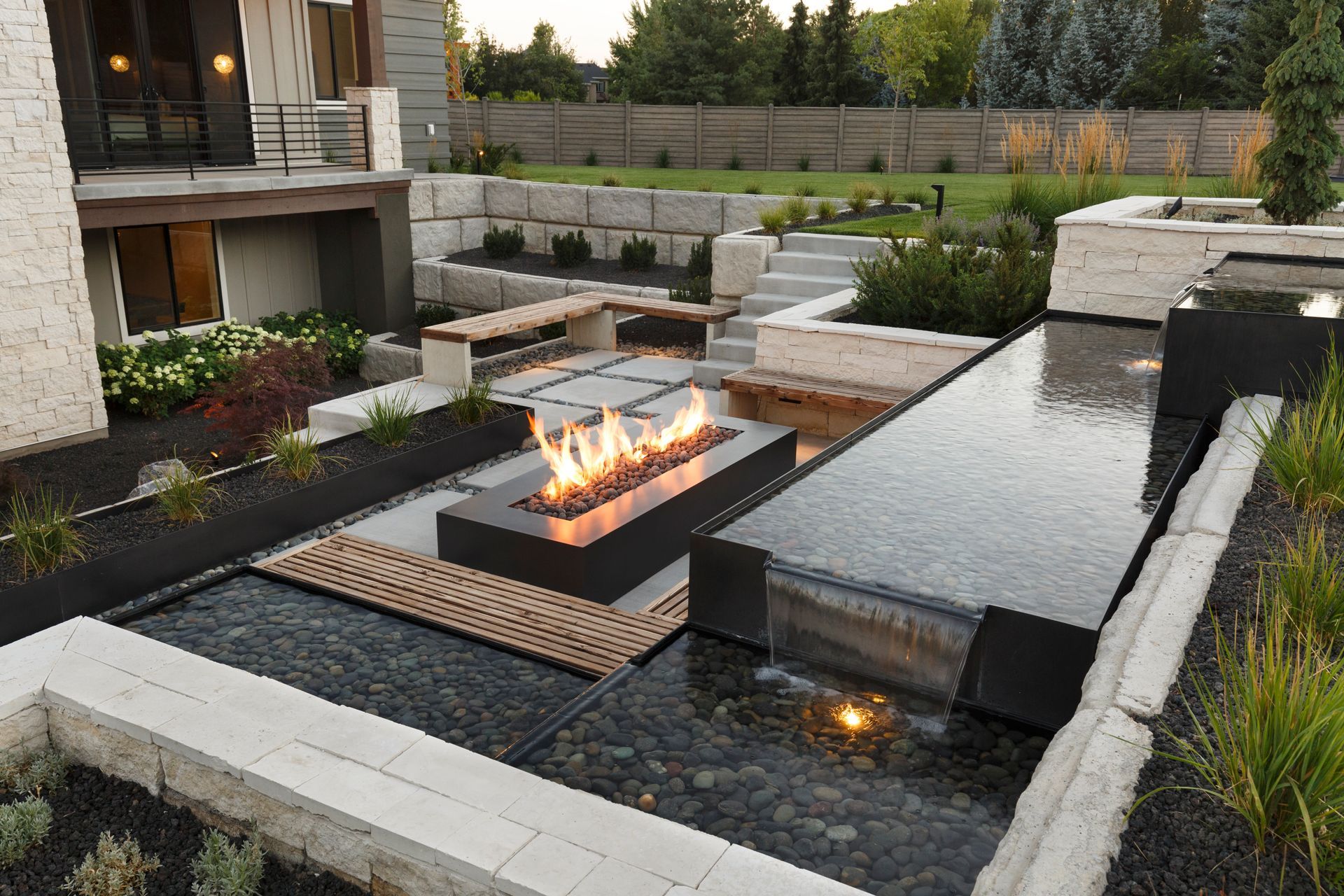 A tiered stone garden with a cascading waterfall, stone steps, and colorful flowers along a landscaped pathway.