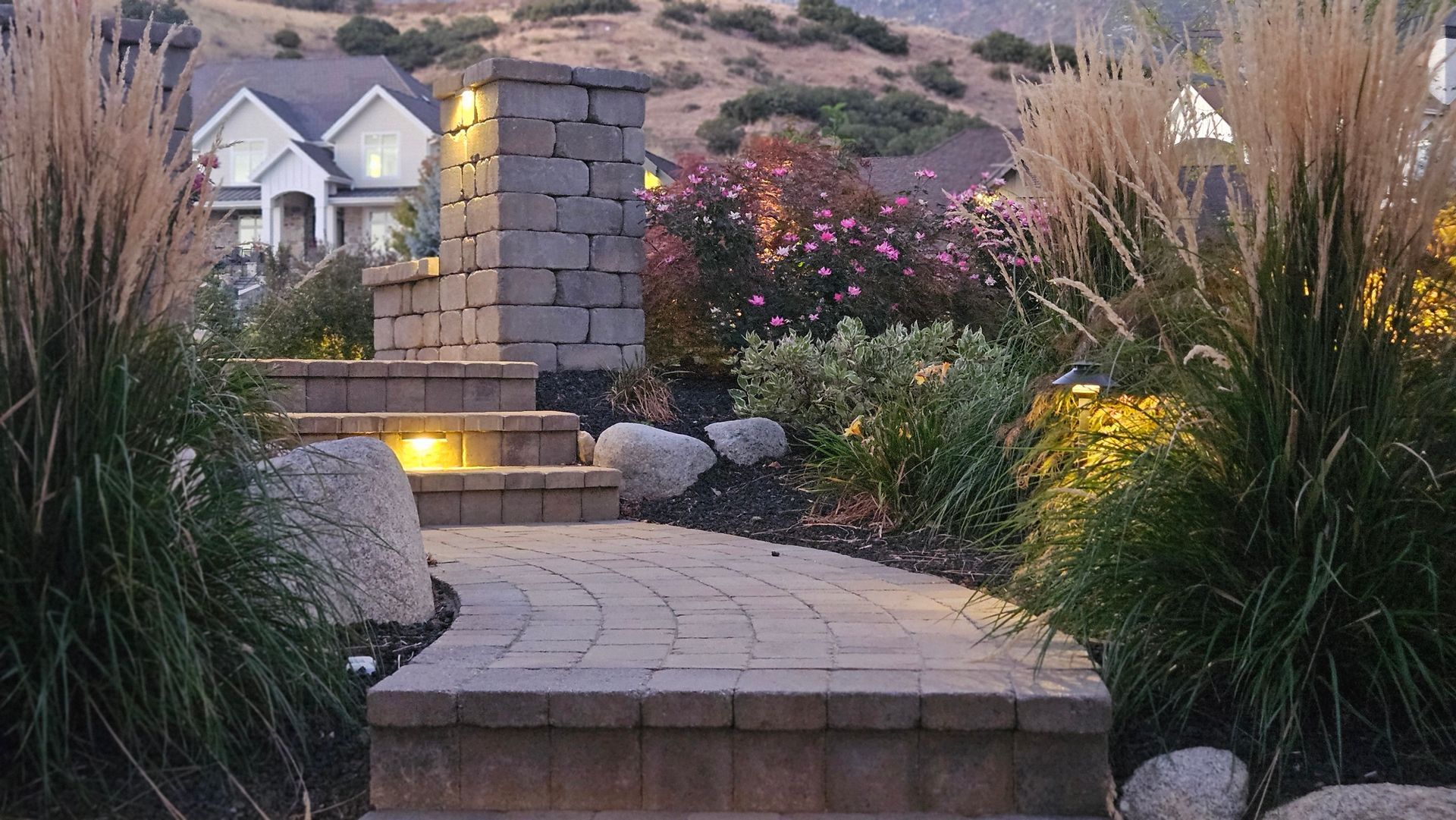 A tiered stone patio with a water feature, lounge chair, gas fire, and landscaped flower pots at dusk.