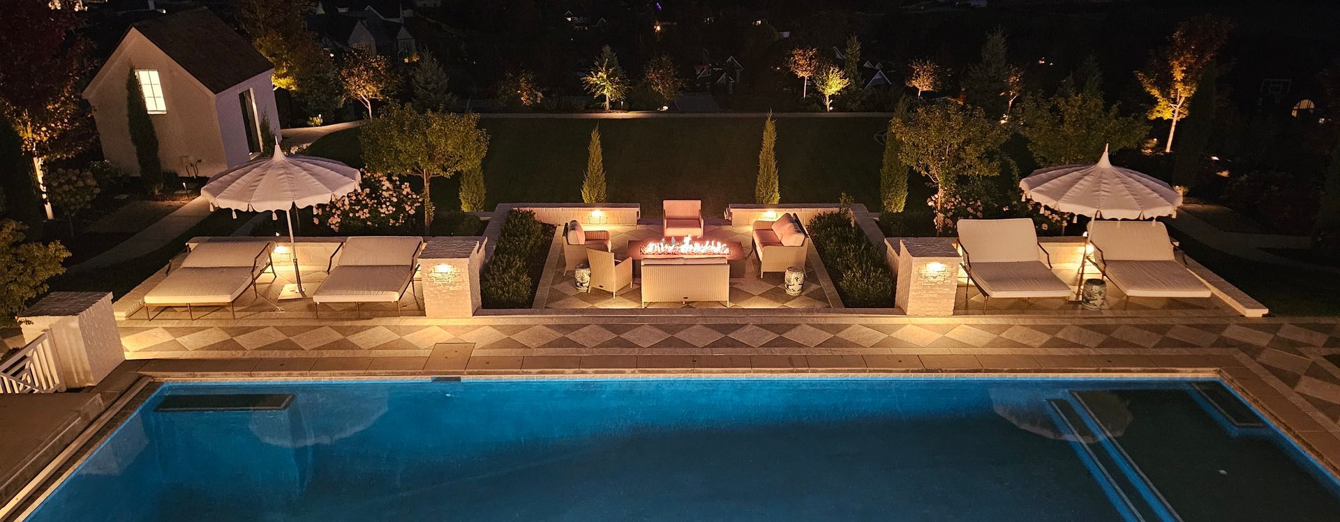 A tiered stone patio with a water feature, lounge chair, gas fire, and landscaped flower pots at dusk.