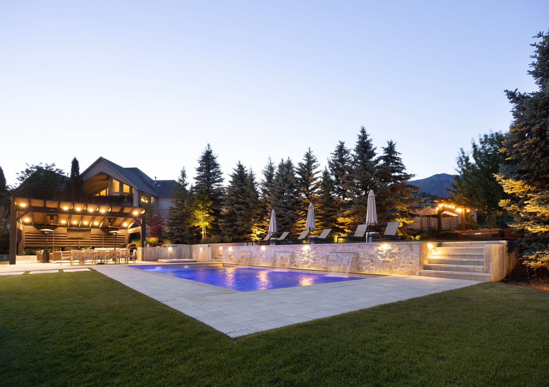 An outdoor kitchen under a stone pavilion, featuring a bar with four chairs, a built-in grill, and evening lighting.