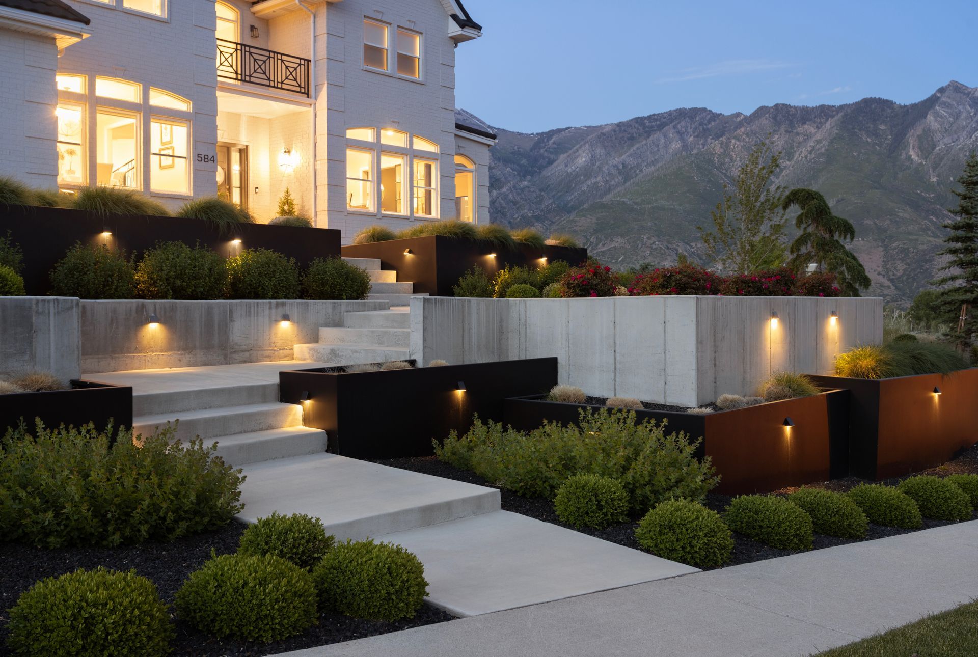 A tiered stone garden with a cascading waterfall, stone steps, and colorful flowers along a landscaped pathway.