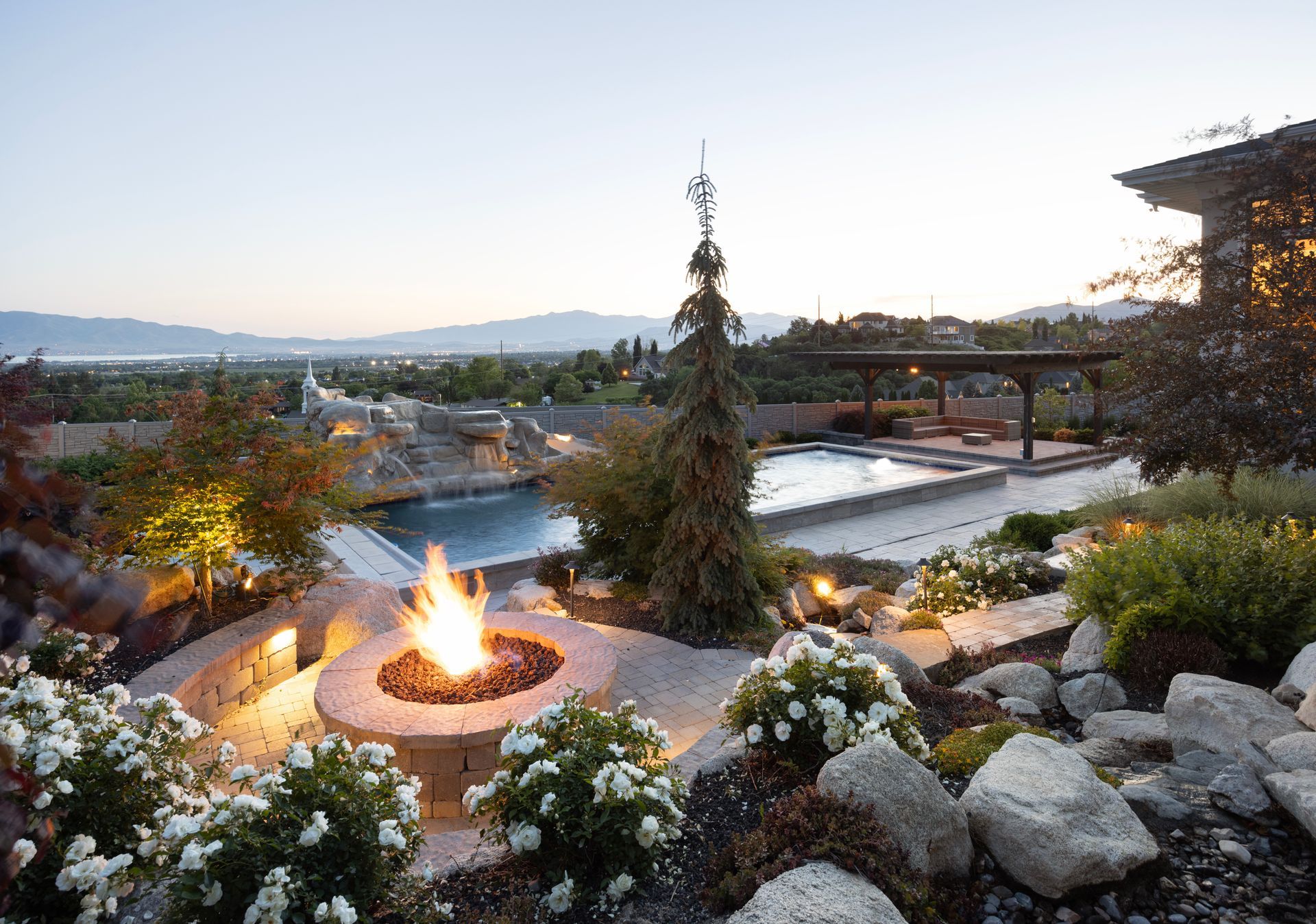 A stone patio with a rectangular swimming pool, a water feature, a waterslide tunnel, and tiered landscaping.