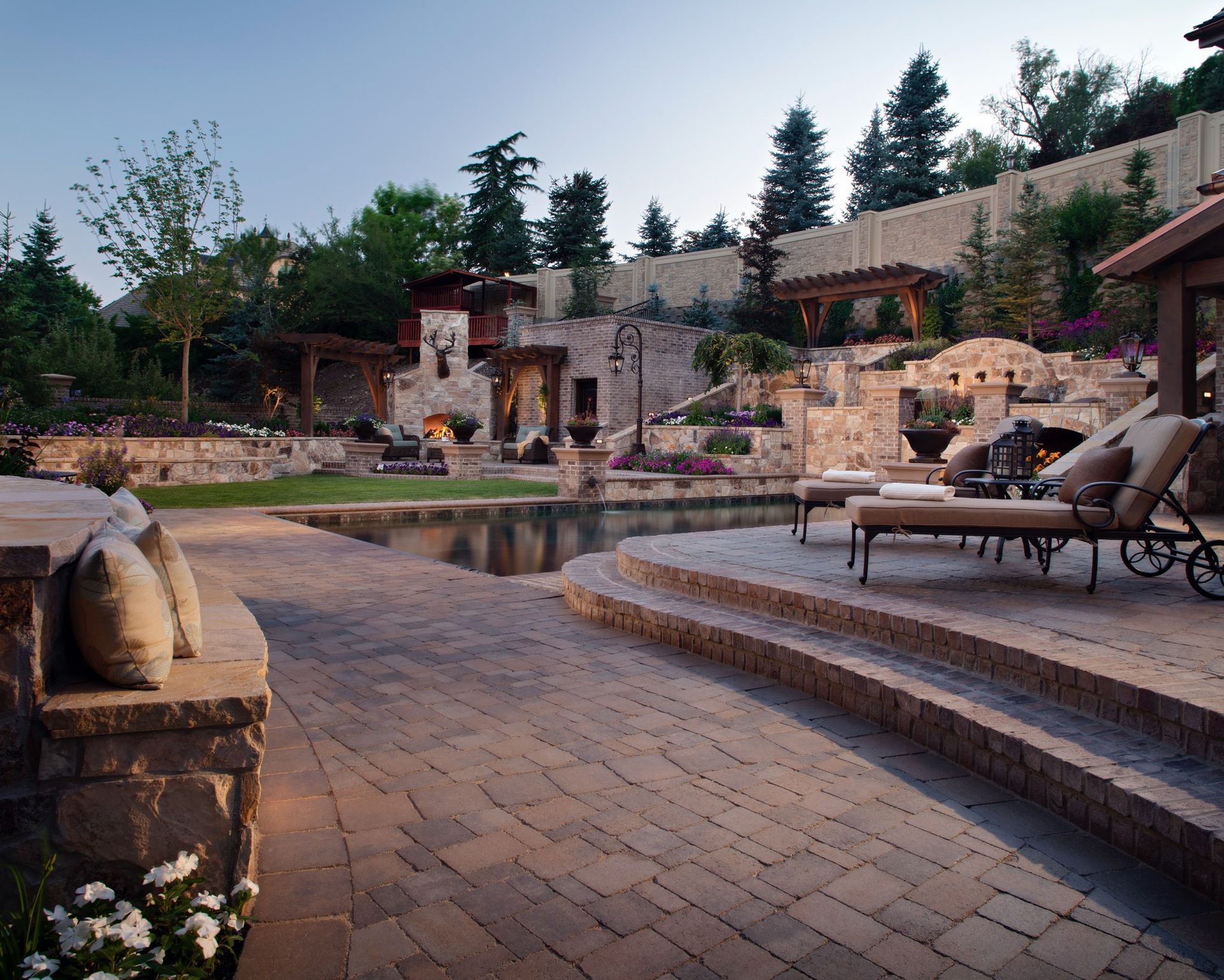 A tiered stone garden with a cascading waterfall, stone steps, and colorful flowers along a landscaped pathway.