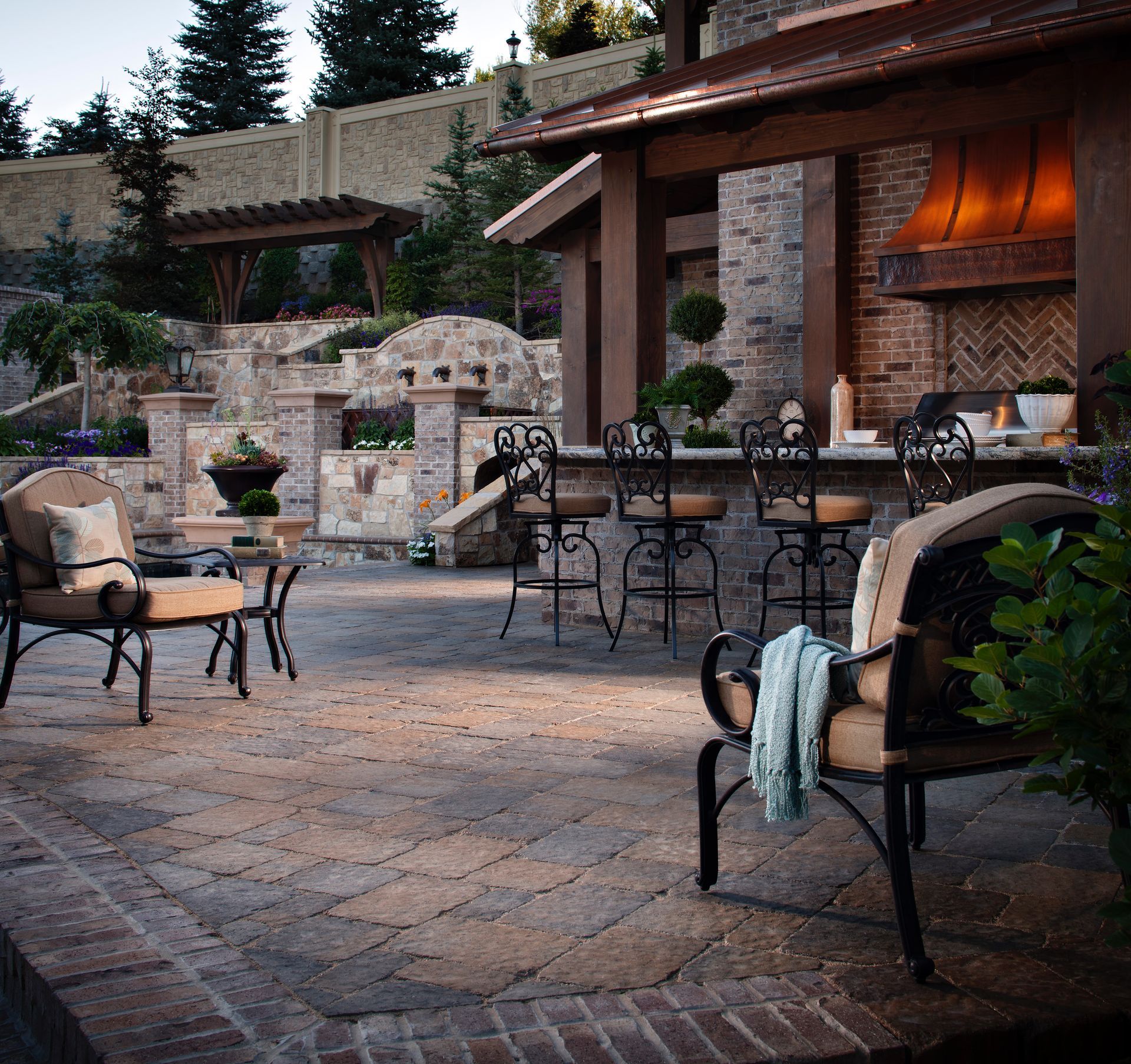 A tiered stone patio with a water feature, lounge chair, gas fire, and landscaped flower pots at dusk.