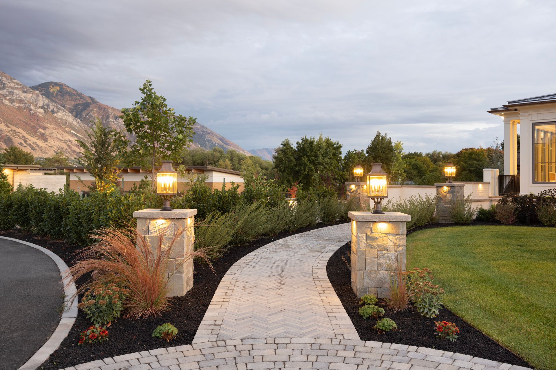 A tiered stone patio with a water feature, lounge chair, gas fire, and landscaped flower pots at dusk.
