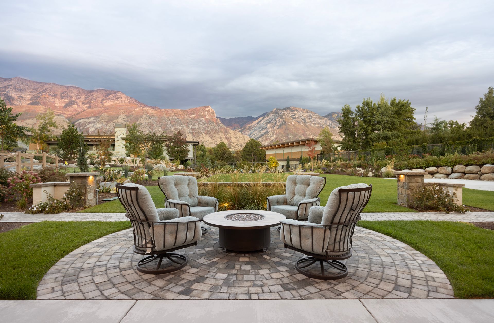 A tiered stone patio with a water feature, lounge chair, gas fire, and landscaped flower pots at dusk.