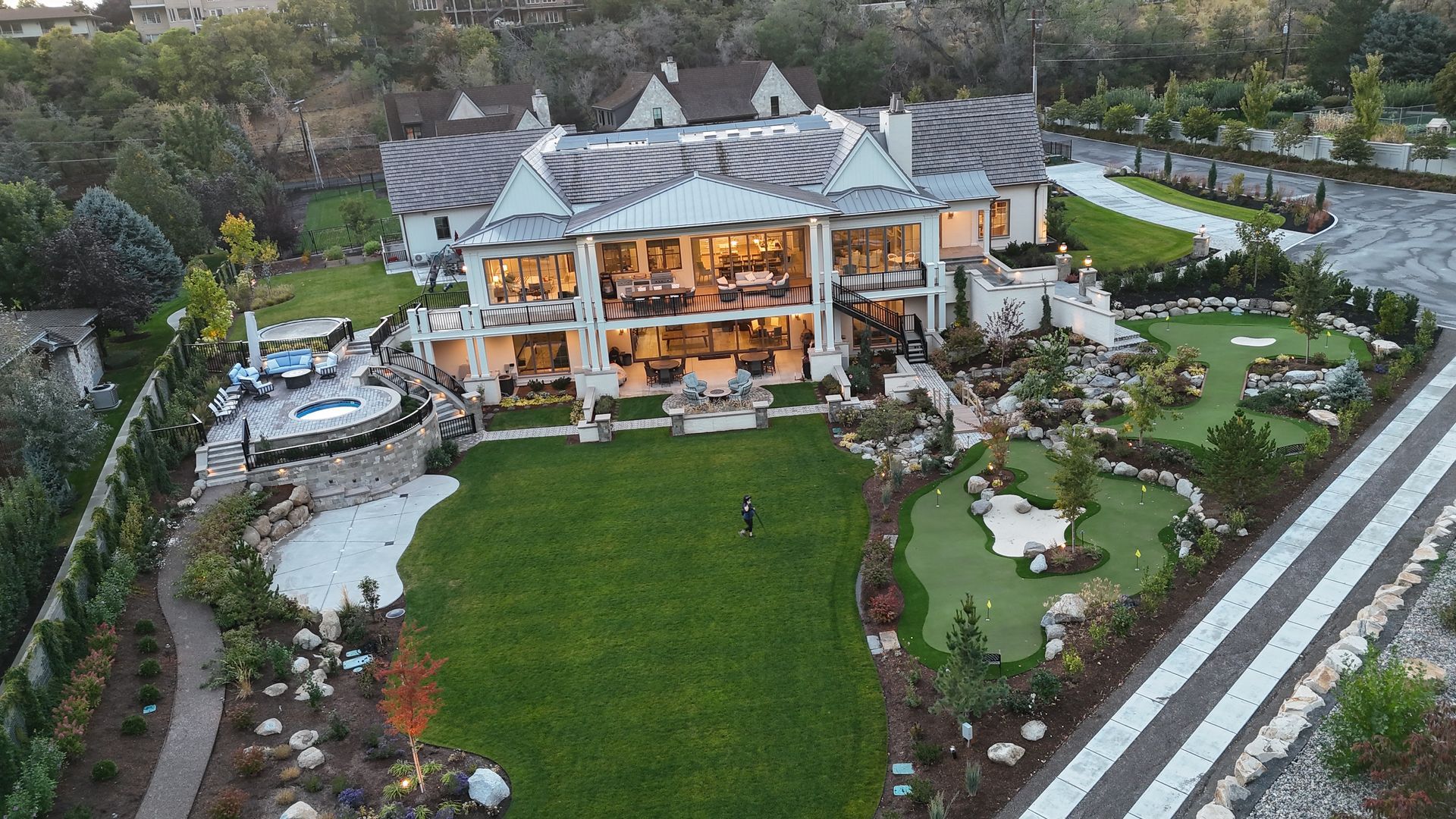 A tiered stone patio with a water feature, lounge chair, gas fire, and landscaped flower pots at dusk.