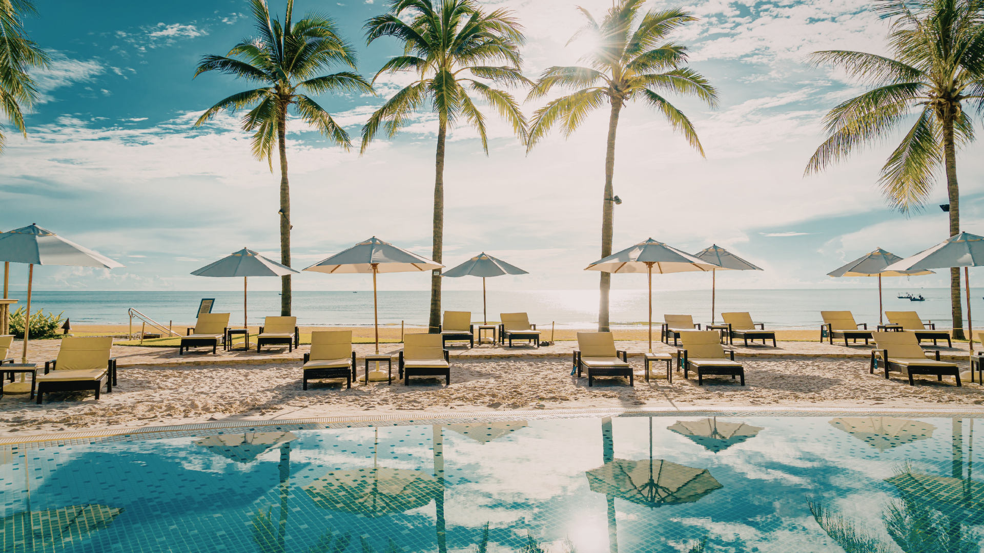 Beach resort with lounge chairs, palm trees, umbrellas, and a turquoise pool beside the ocean at sunset