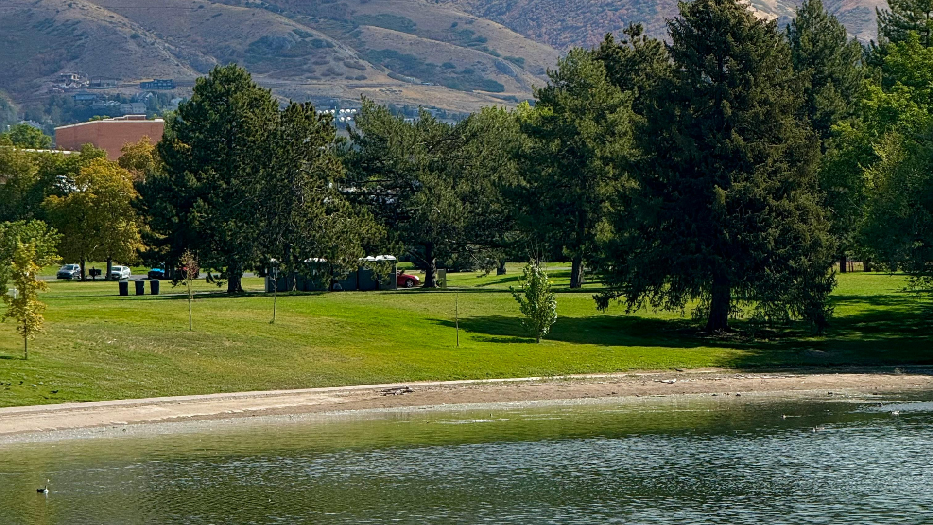 Lakeside park with green lawn, trees, and mountains in the background.