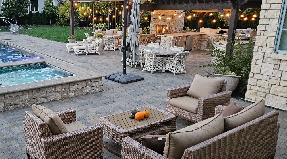 A backyard patio at dusk featuring wicker lounge seating, a coffee table, a hot tub, and an outdoor kitchen area.