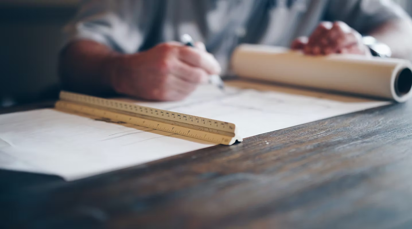 A person working on architectural blueprints with a drafting scale on a wooden table.