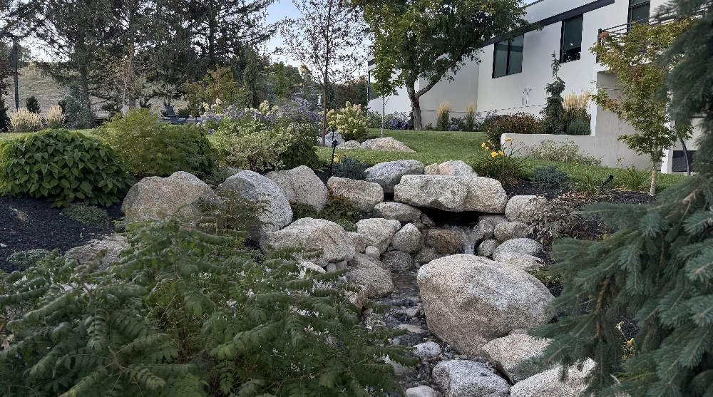 A rock-lined garden stream flows through a landscaped yard with green bushes and trees near a white building.