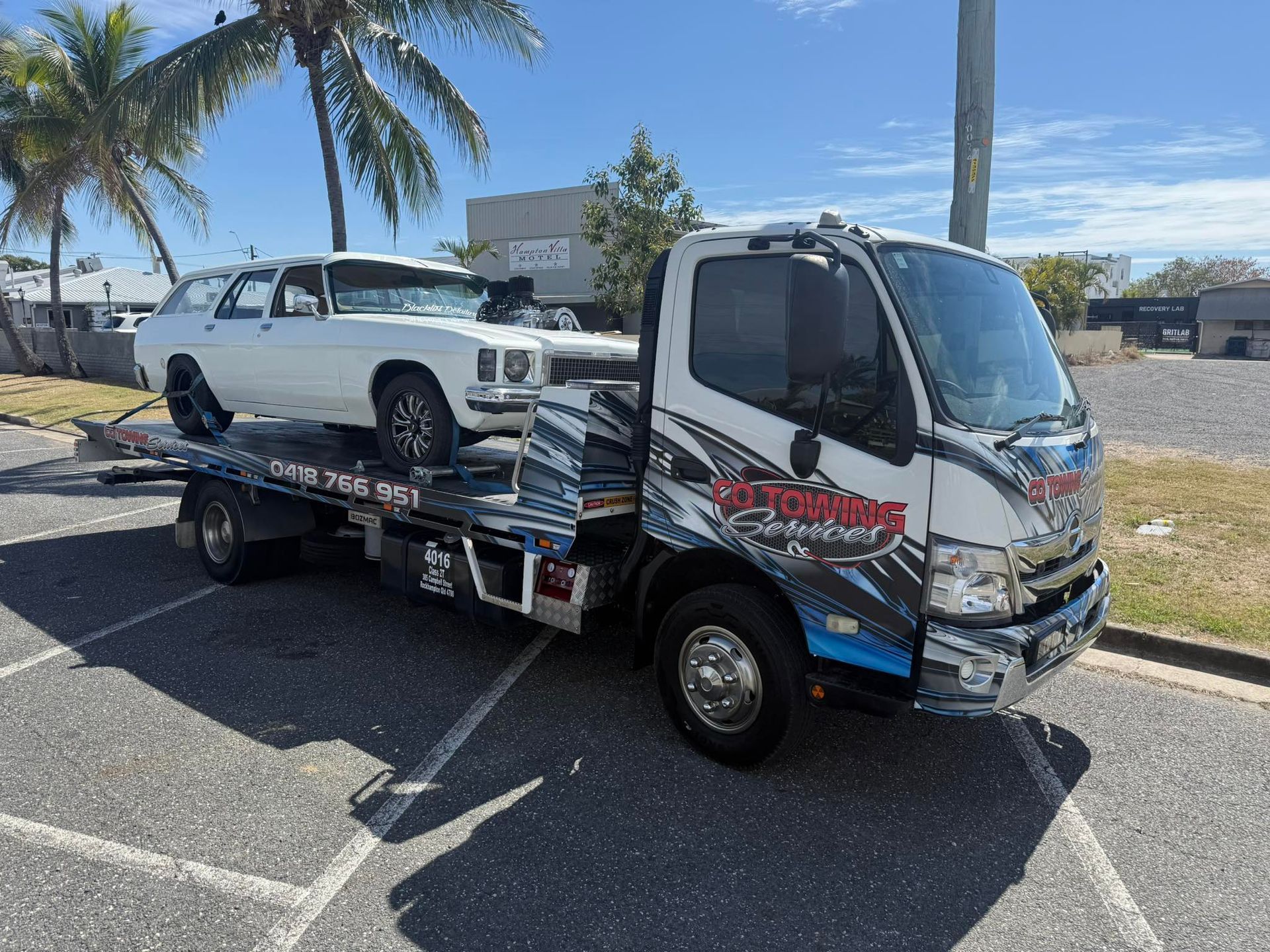 White car on a tow truck, parked on pavement. Sunny day with palm trees.