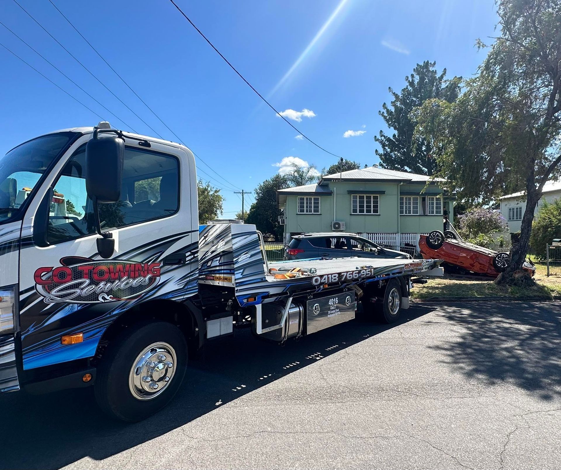 Tow truck with a damaged car on the flatbed, near a house on a sunny day.