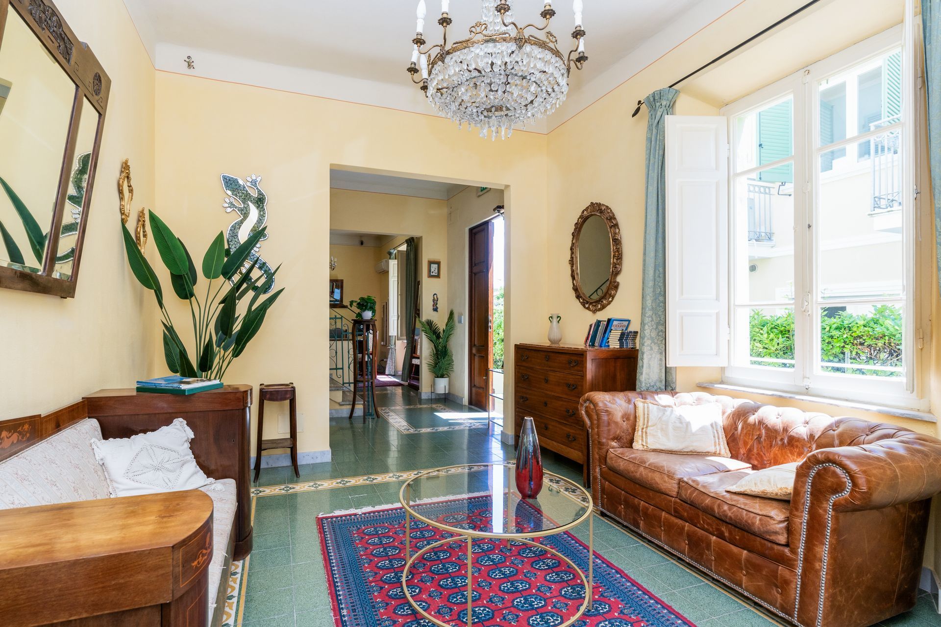 Living room with a leather couch, plants, a rug, and a chandelier; light yellow walls and a view through a doorway.