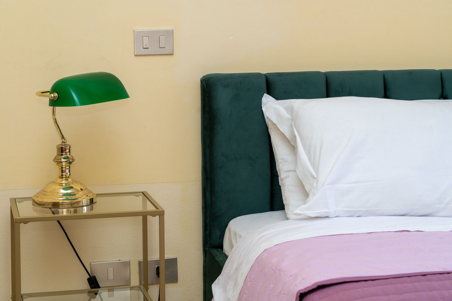 Green lamp on gold bedside table next to a bed with green headboard and pink blanket.