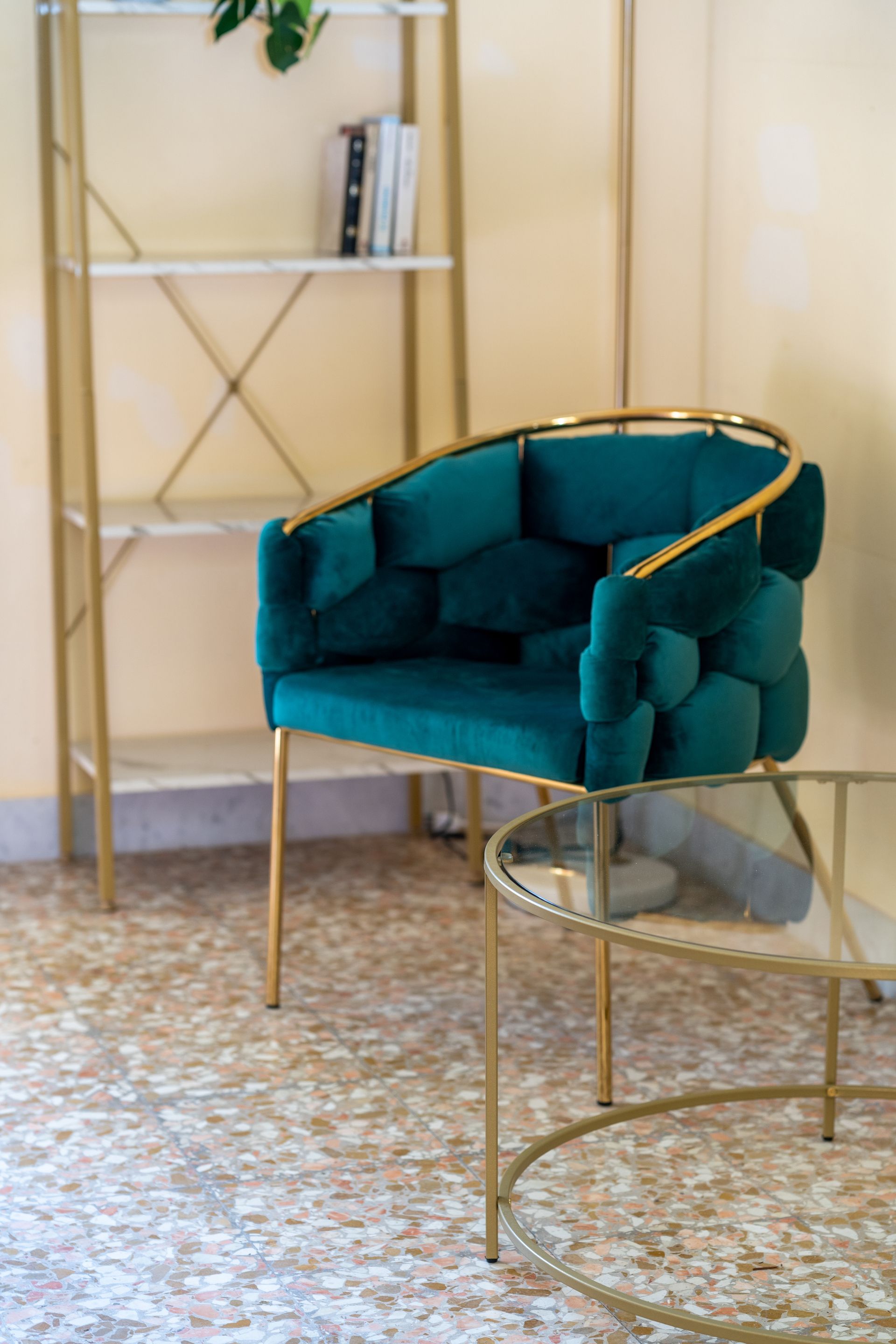 Teal velvet armchair with gold frame, marble table, bookshelf in a room with terrazzo flooring.