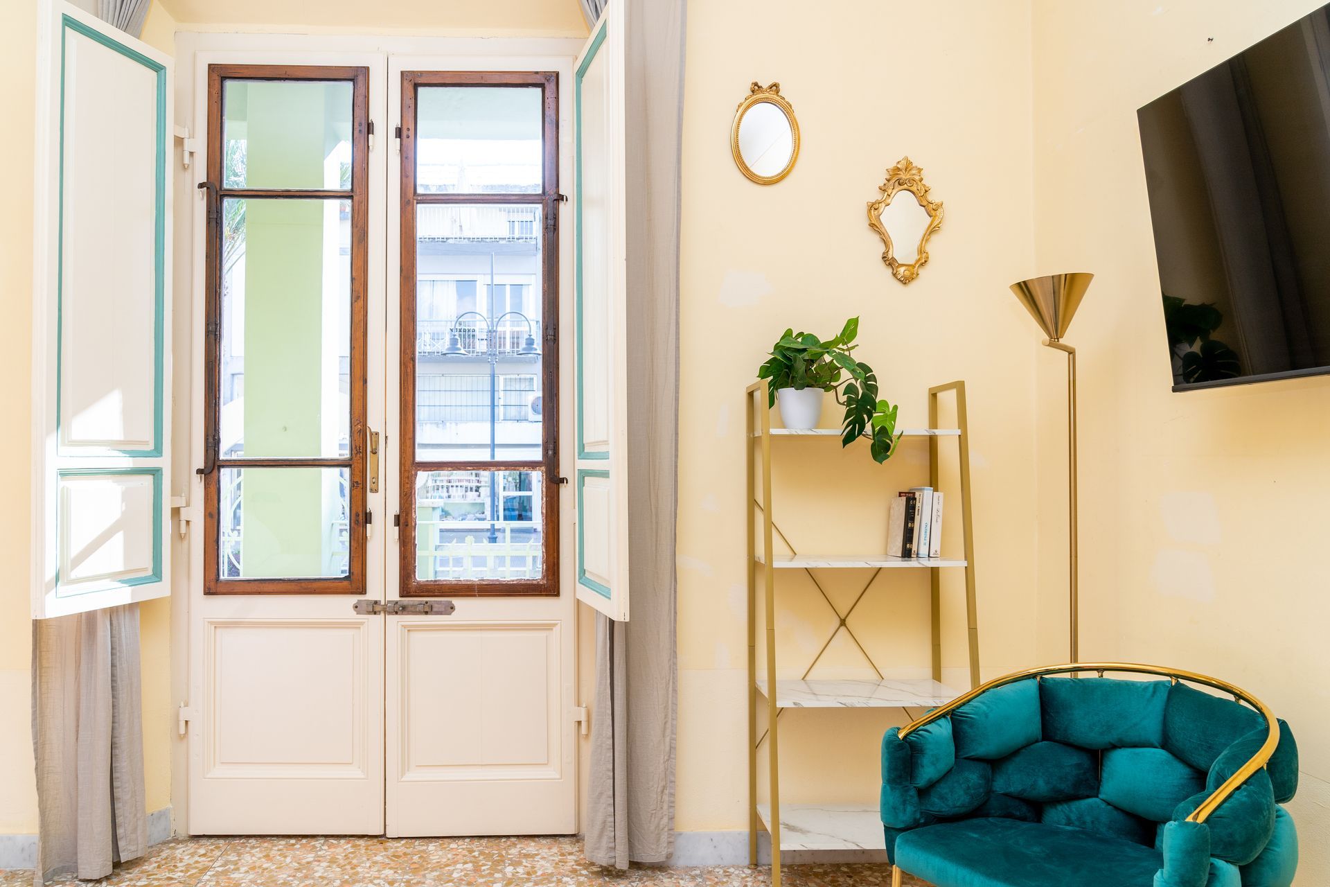 Interior with teal chair, gold shelving unit with a plant, open door, and a wall-mounted TV.