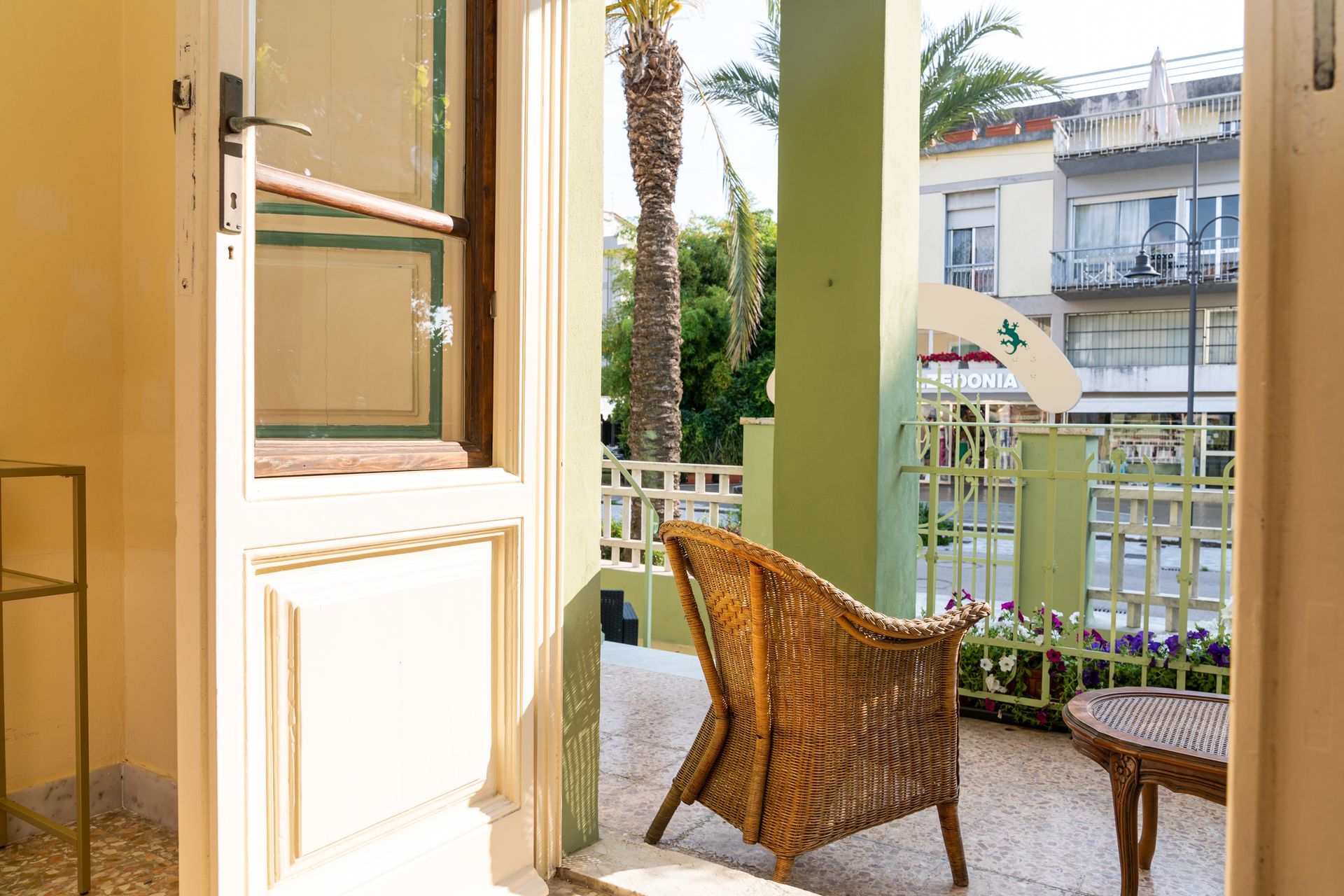 Open door leads to a balcony with wicker chair and plants. Palm tree and building visible.