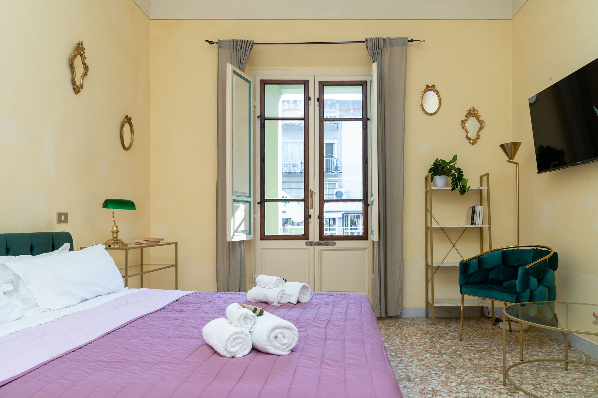 Bedroom with open window, bed, velvet chair, and decorative mirrors on a yellow wall.