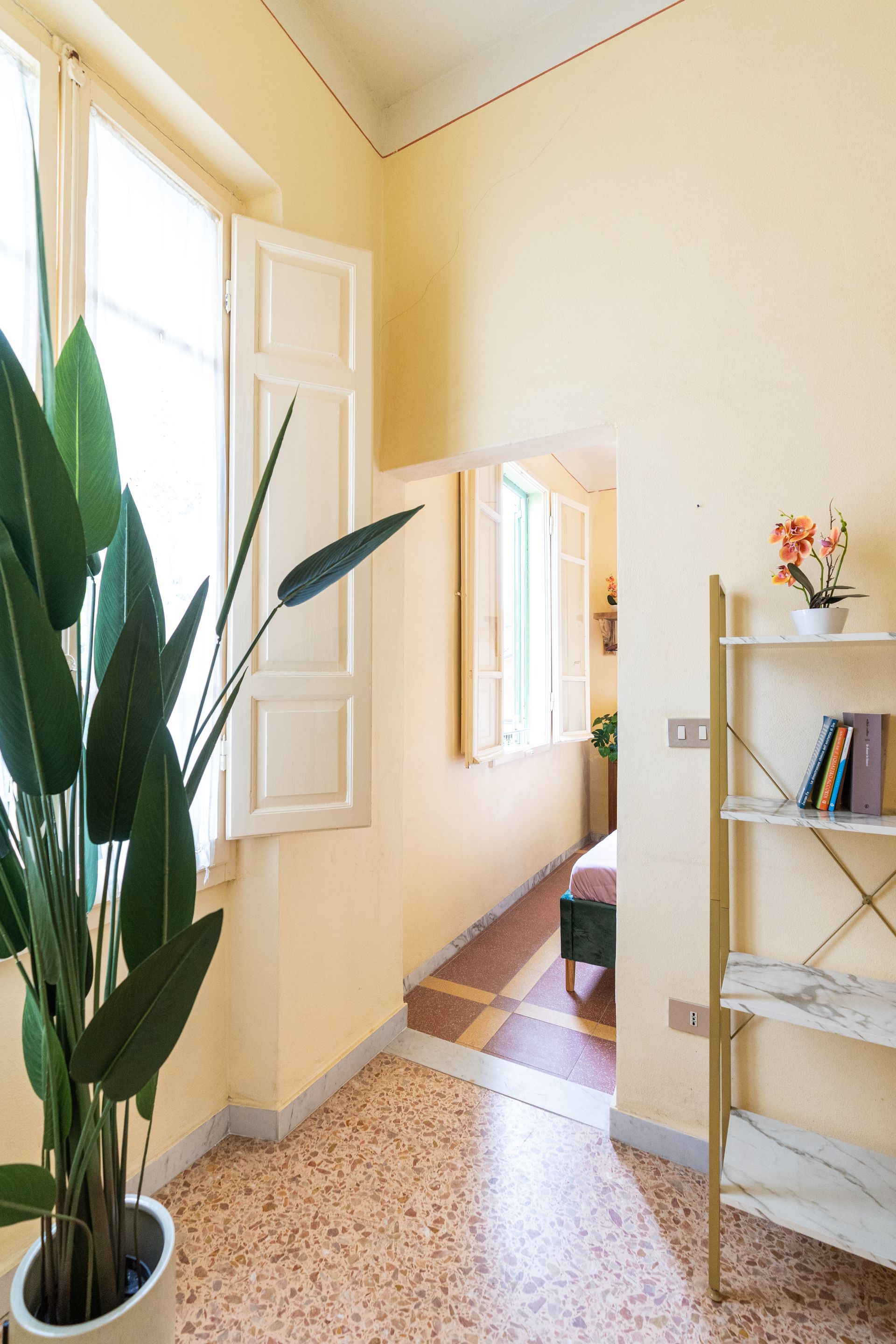 Interior view of a light-yellow hallway with a window, plant, and a bookshelf; doorway leads to a bedroom.