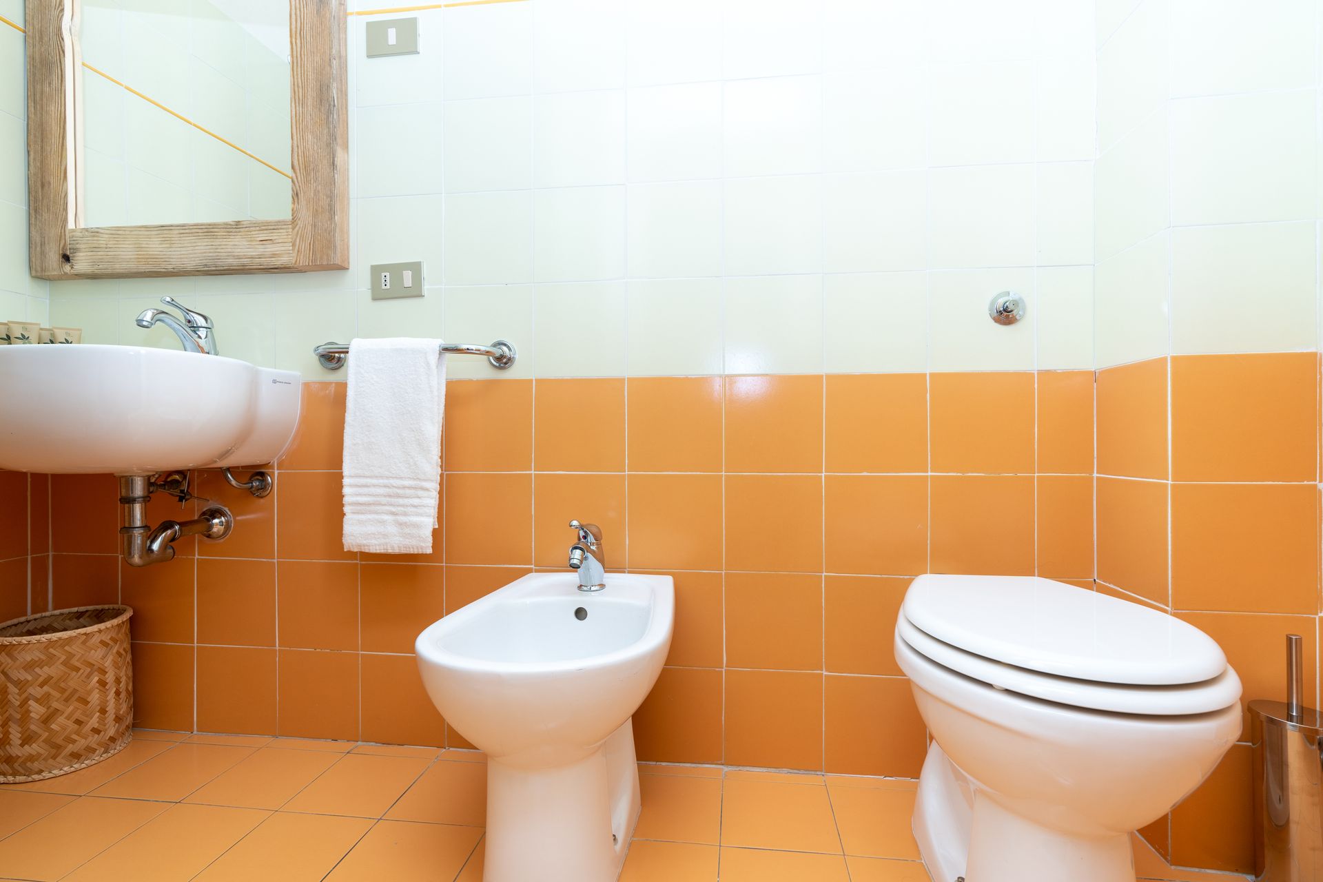 Bathroom with orange tiled walls, sink, bidet, and toilet. A mirror hangs above the sink.
