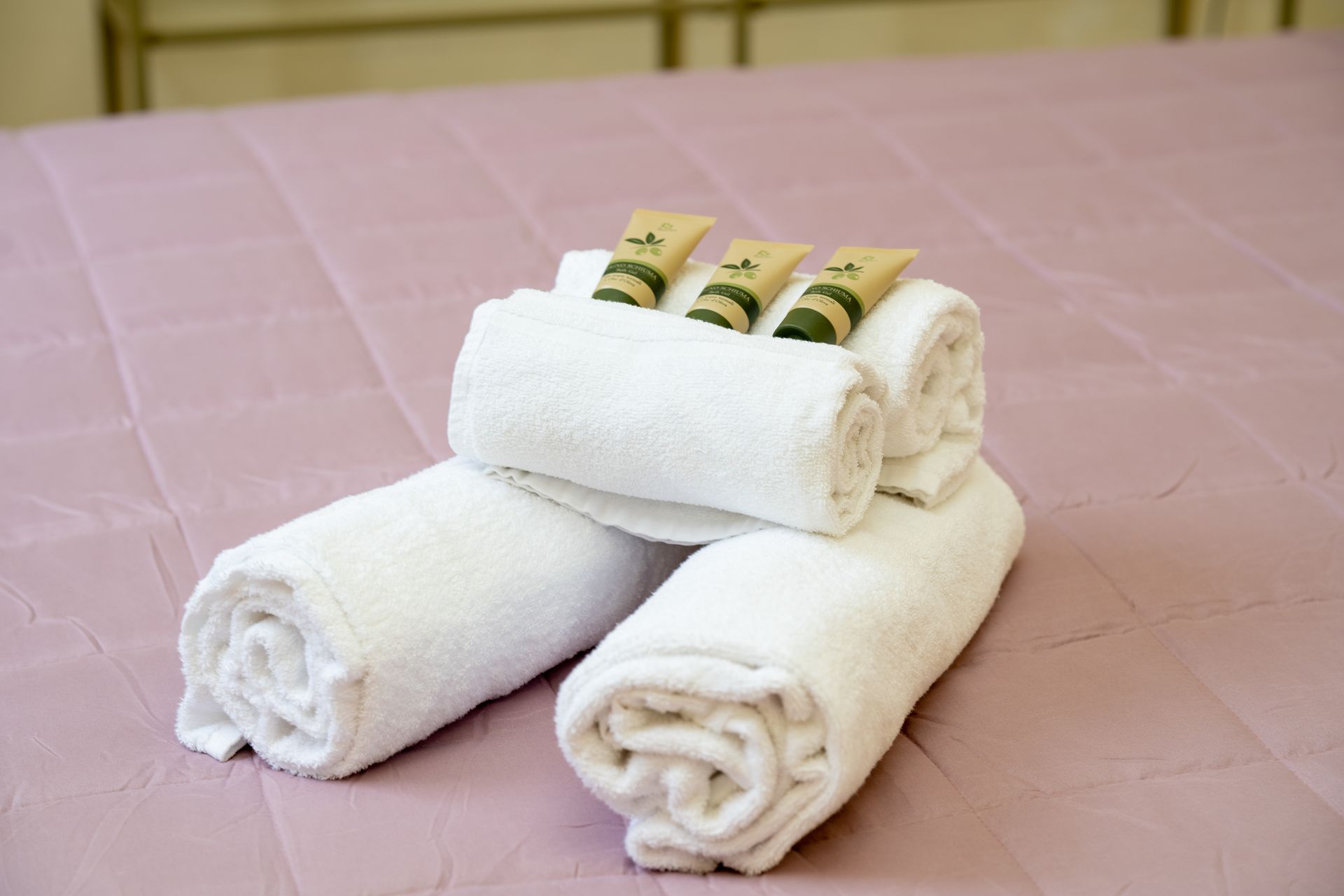 Rolled white towels on a pink bed with three small toiletries on top.