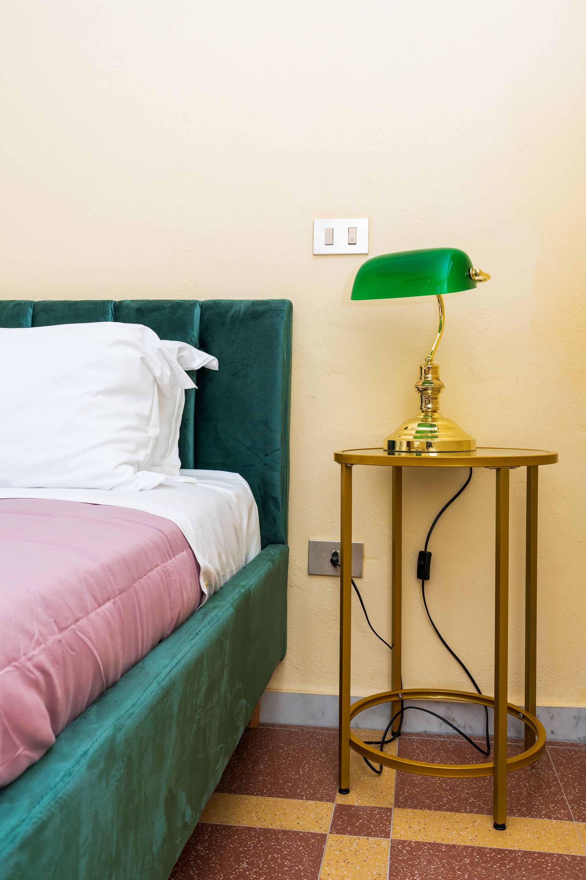 Teal bed with pink blanket, gold side table with green lamp. Tan wall, checkered floor.
