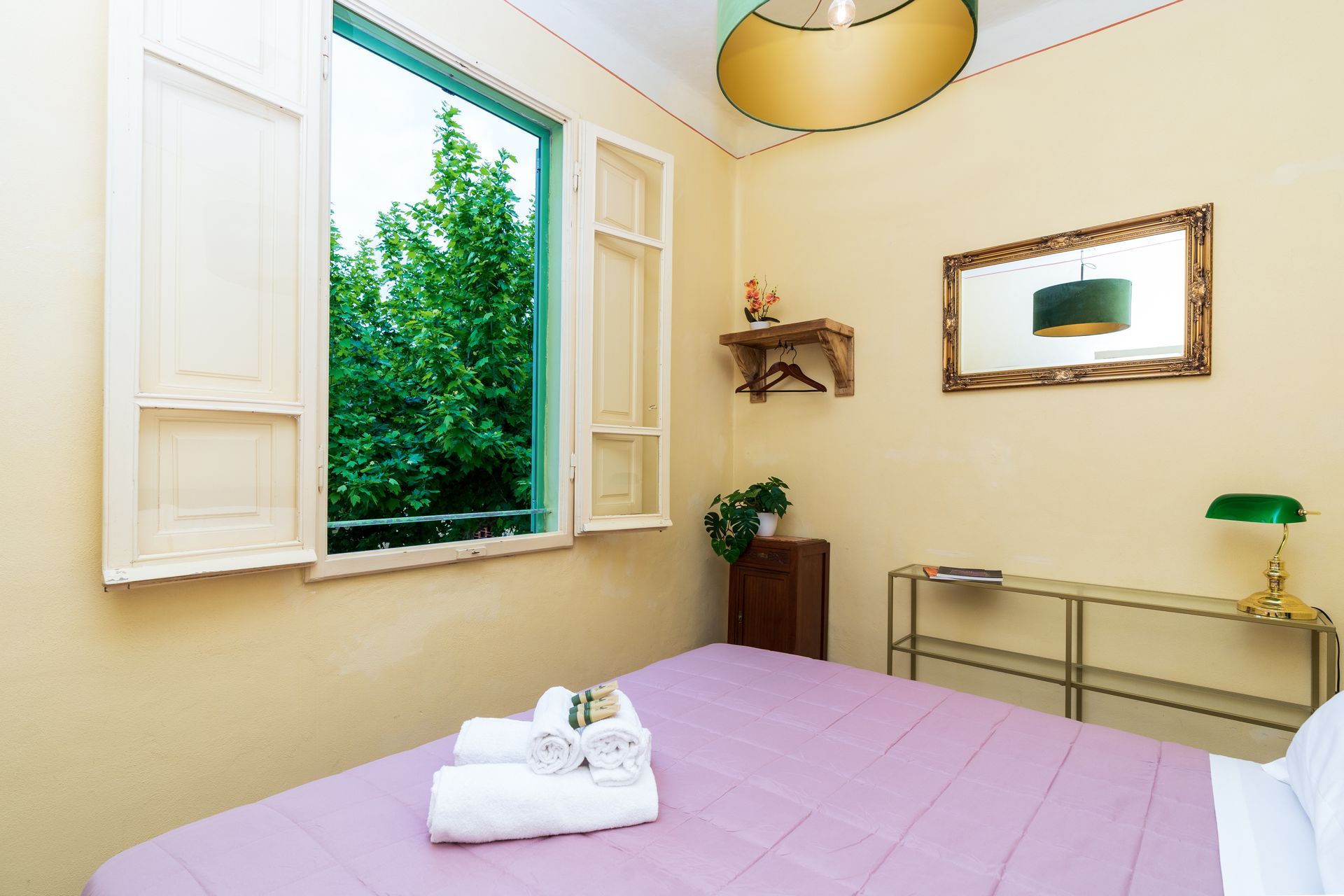 Bedroom with open window, bed, and decorative items. Pale yellow walls, green tree outside.