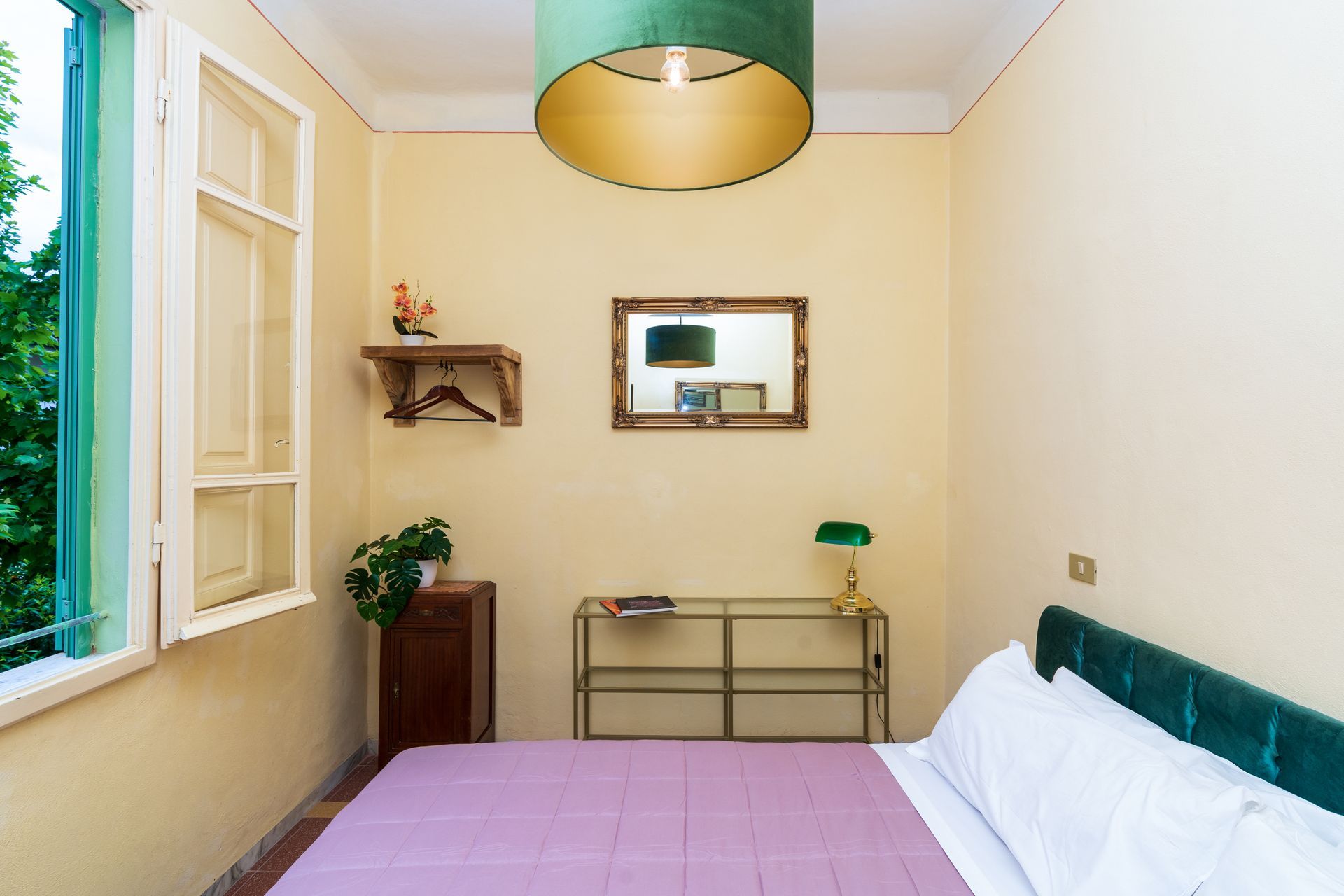 Bedroom with green velvet headboard, pink bedspread, window, gold-framed mirror, and a green lampshade.