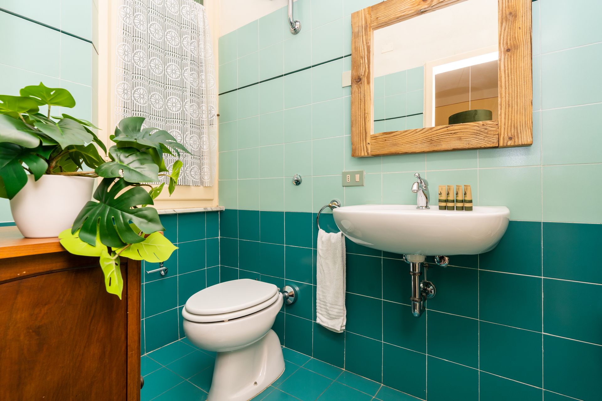 Bathroom with turquoise tile, a round sink, toilet, and wooden-framed mirror. A potted plant sits on a wood cabinet.