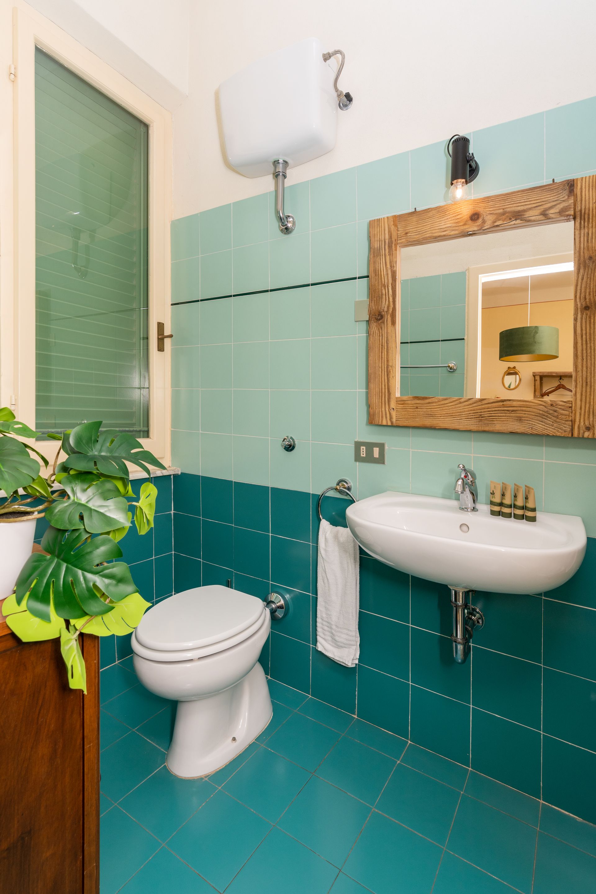 Bathroom with teal tiled walls and floor, white toilet and sink, wooden mirror frame, and plant.