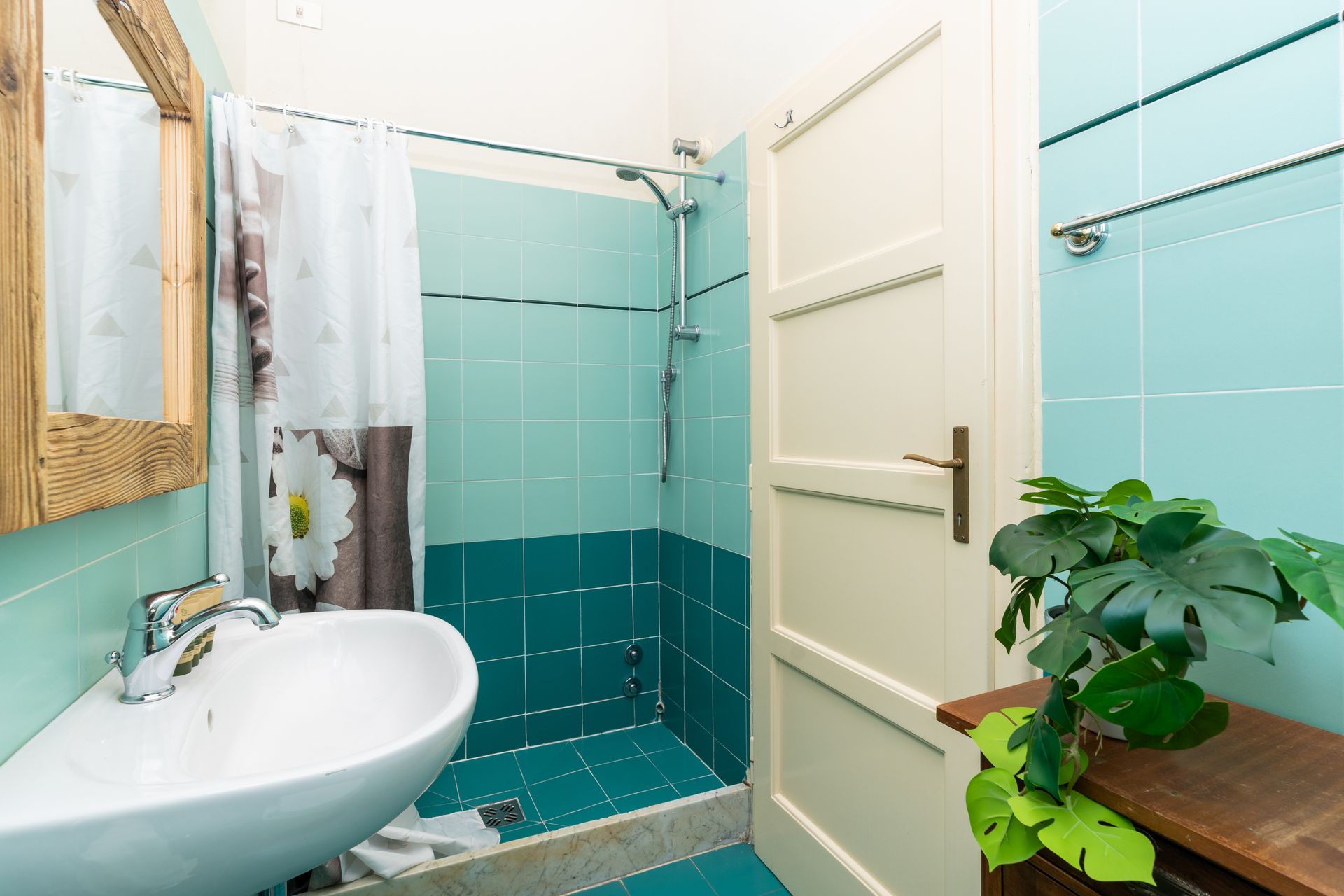Small bathroom with teal tiles, white sink, and wooden-framed mirror. Shower, door, and plant are present.