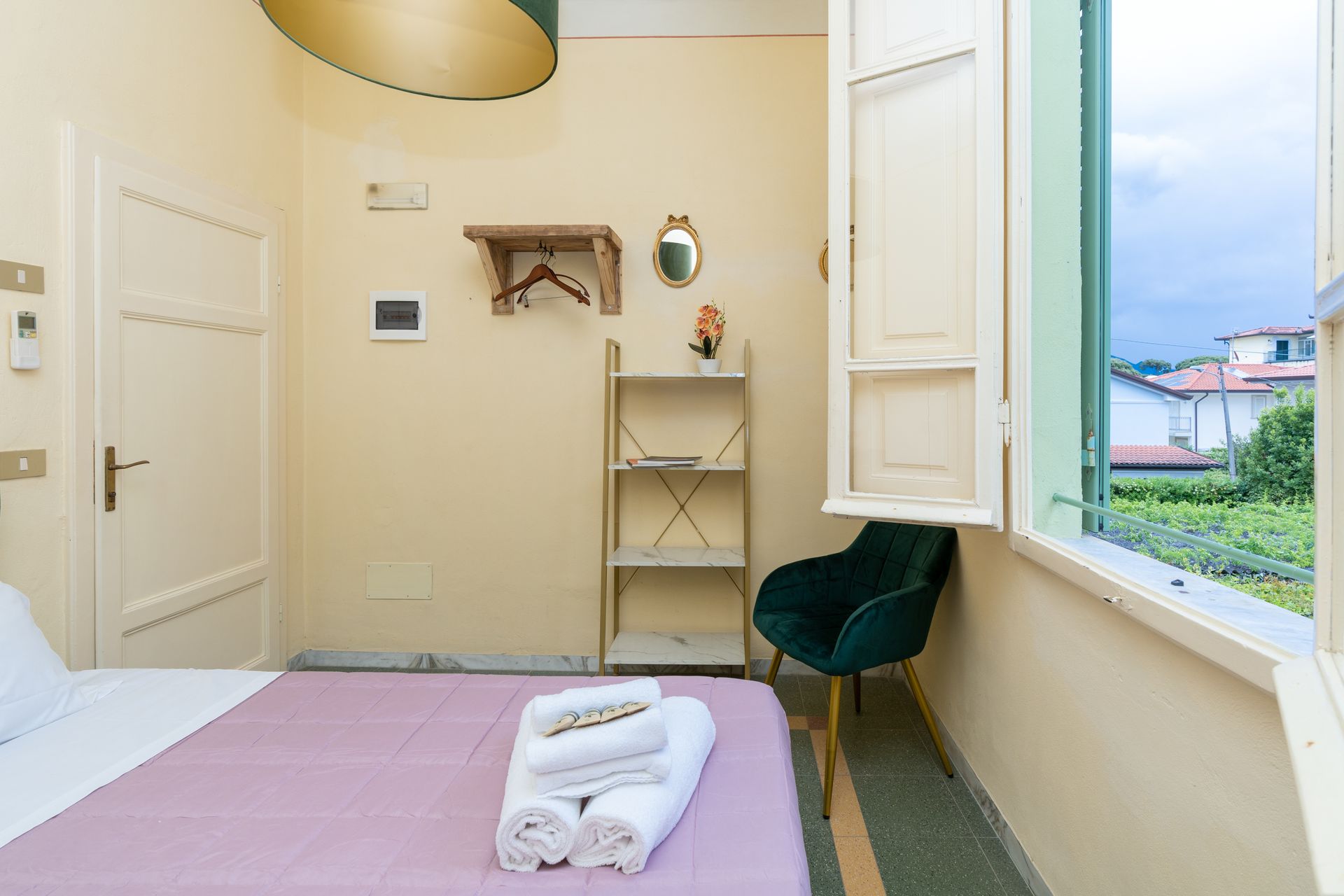Bedroom with open window, bed, shelves, chair, and towels. Green chair, pink bedspread, pale yellow walls.