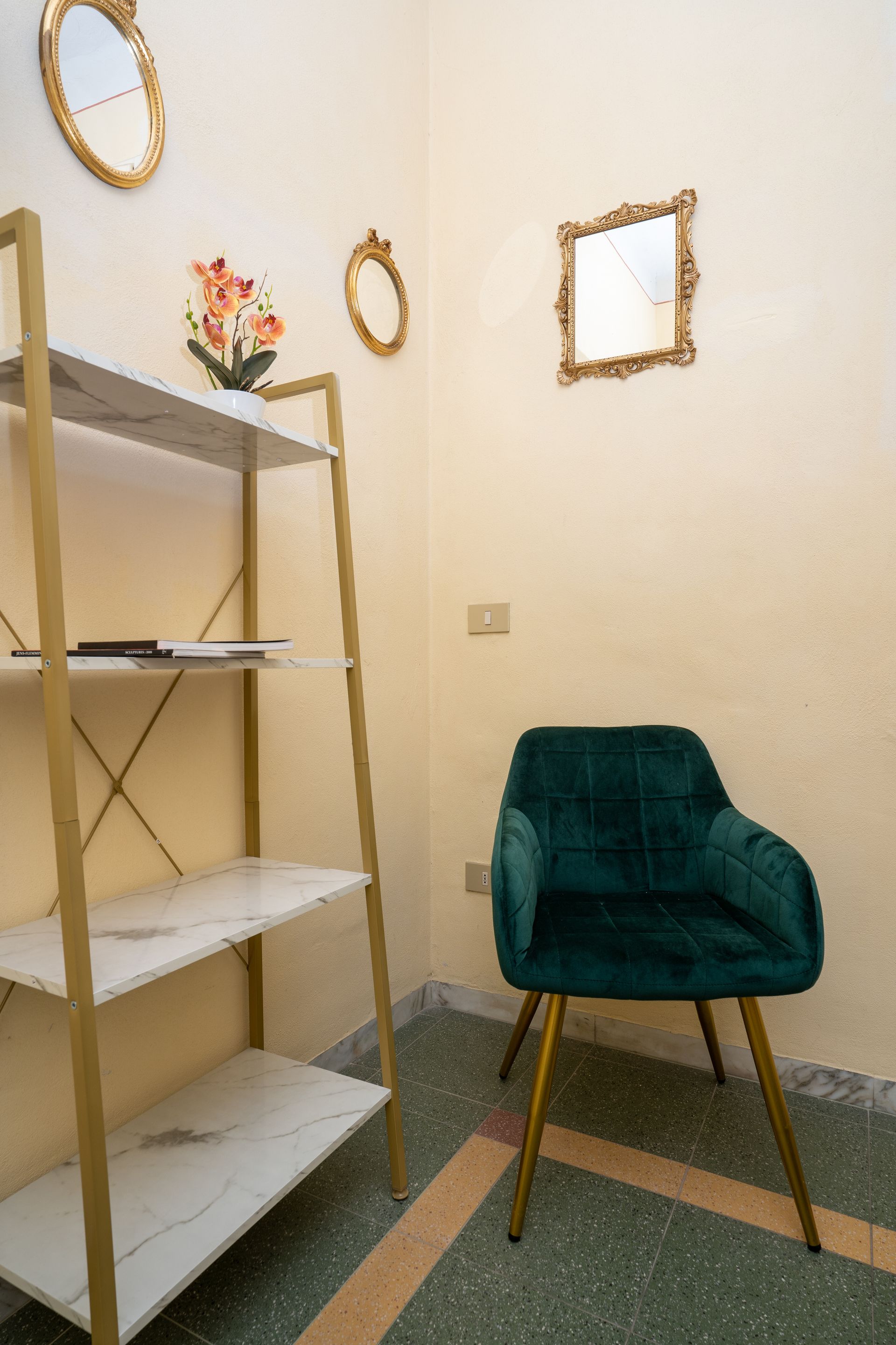 A teal armchair sits near a gold-framed shelving unit and mirrors in a cream-colored room.
