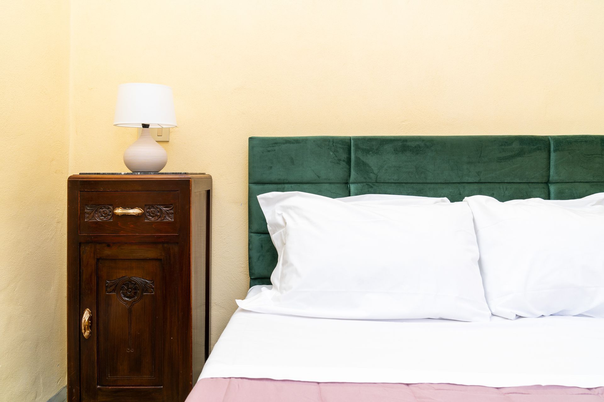 Bedroom with a dark wooden bedside cabinet, lamp, and green headboard with white bedding.