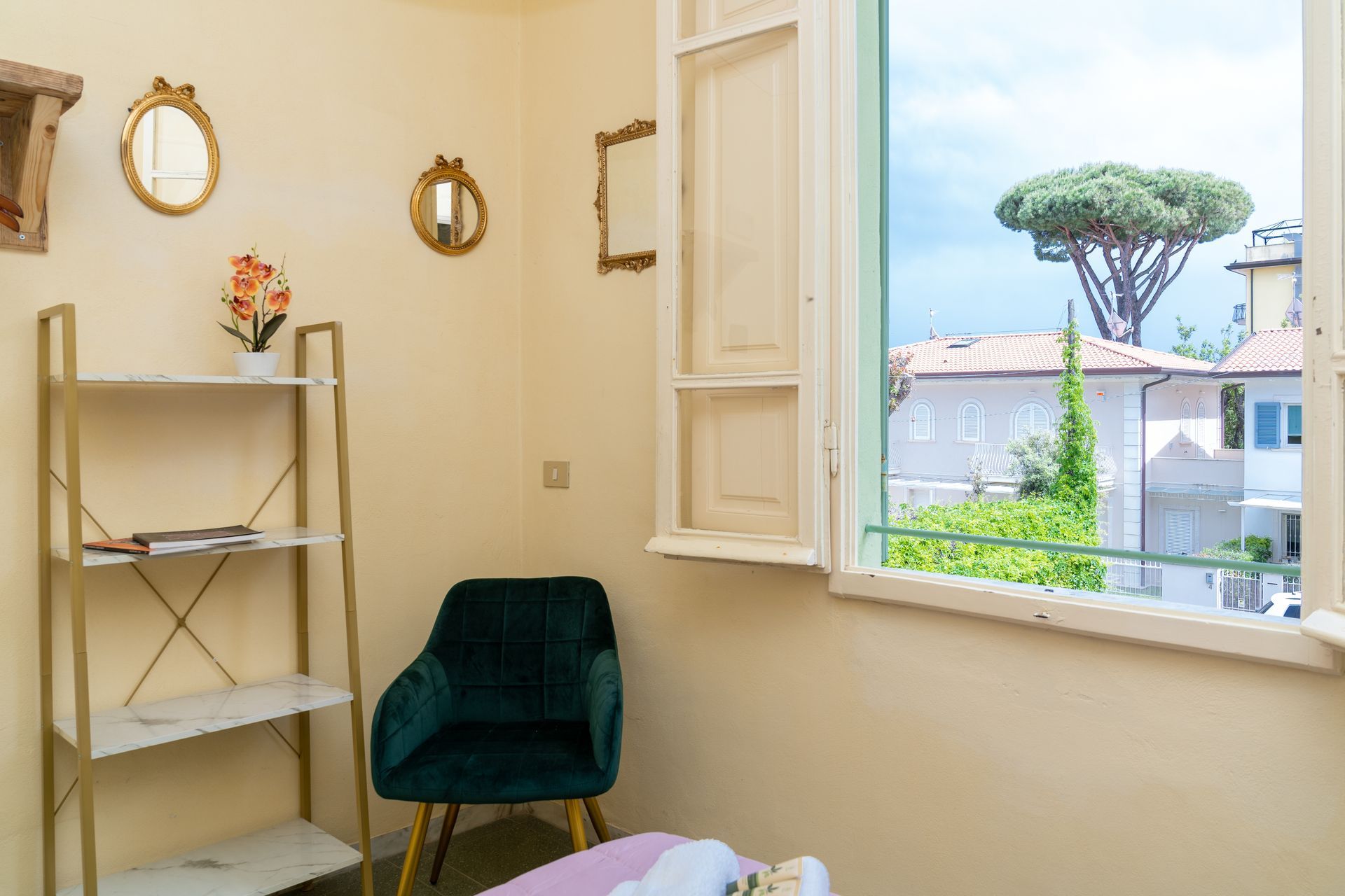 Cozy room with velvet chair, shelving unit, mirrors, and open window with a view of buildings and a tree.