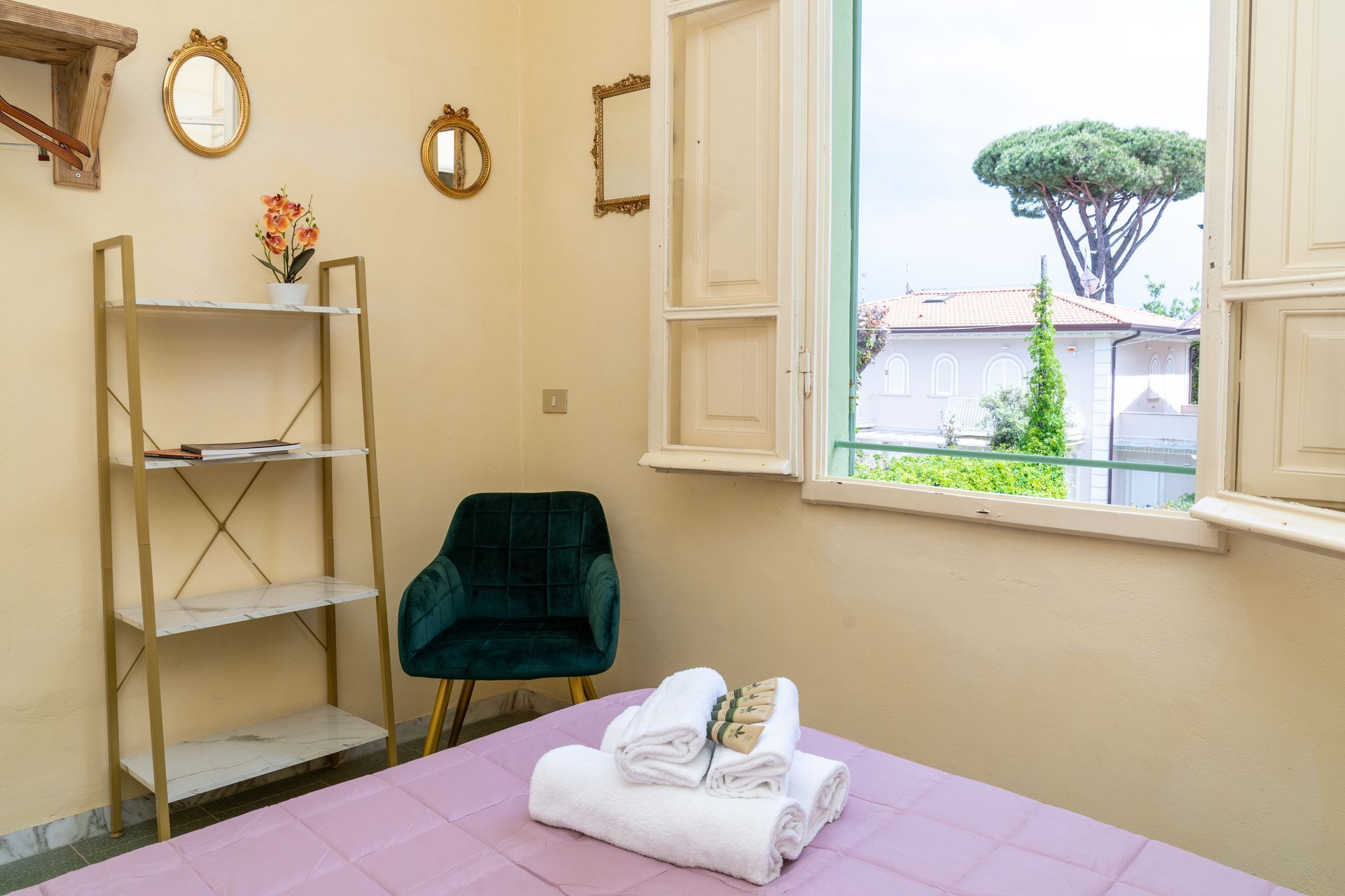 Bedroom with bed, shelf, armchair, window with a view. Light yellow walls, gold accents, green tree visible.