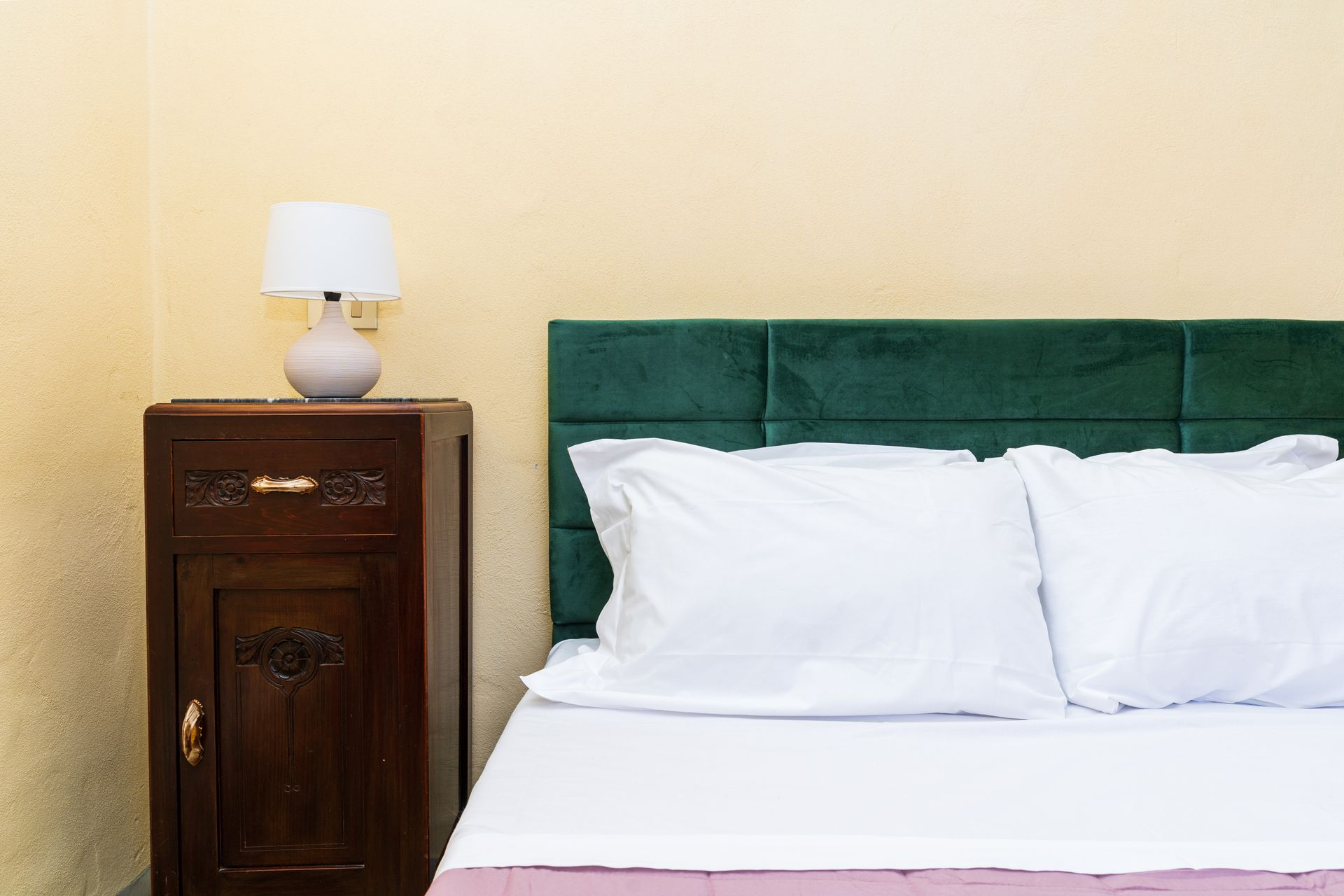 Bedroom with a bed and nightstand. Green headboard, white bedding, and a lamp on the dark wood nightstand.