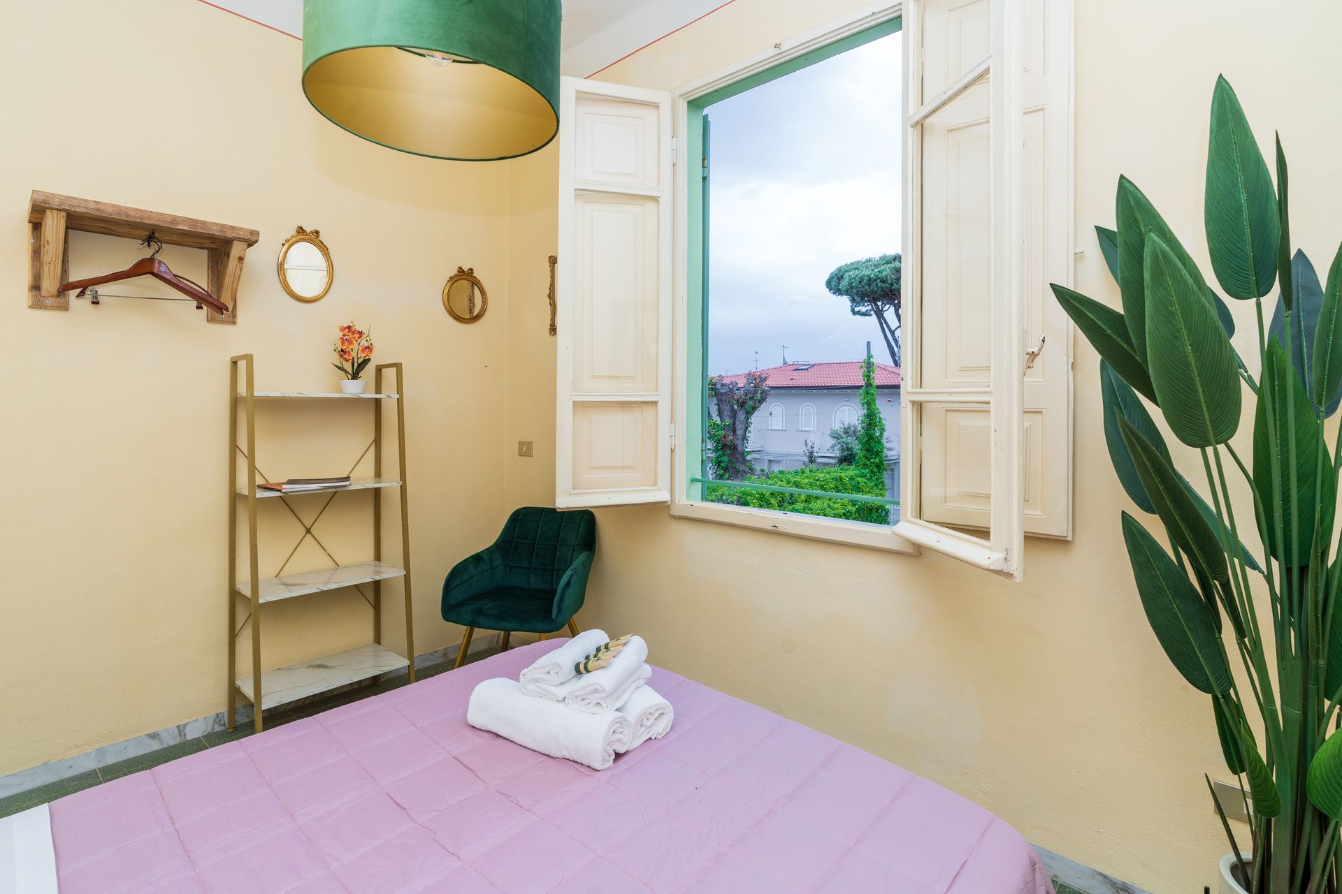 Bedroom with bed, open window, small bookshelf, velvet chair, and decorative plant.
